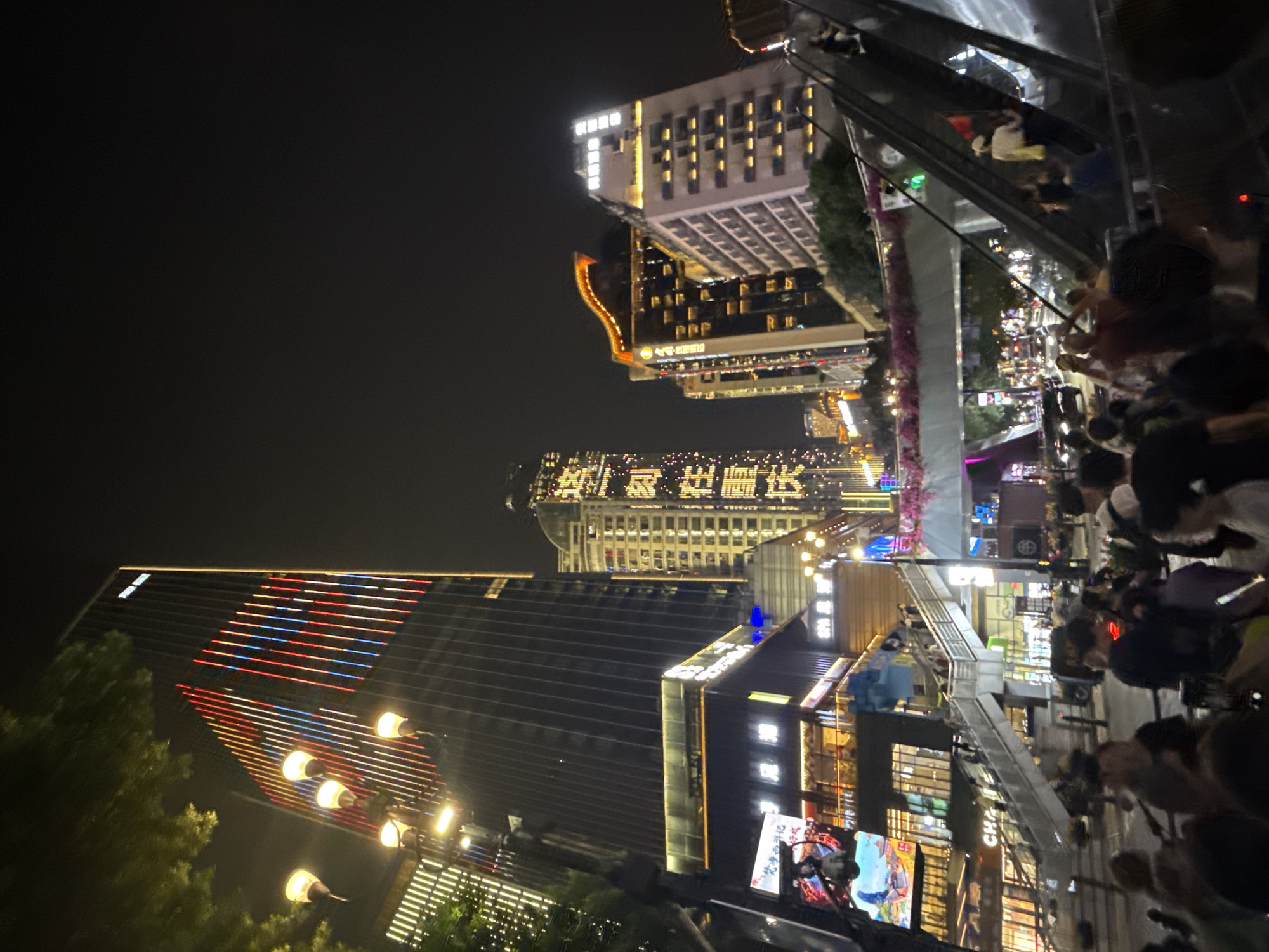 This image captures a bustling night scene in Yuzhong District, China, likely a commercial area with tall buildings and pedestrian traffic. A prominent, illuminated skyscraper dominates the background, its facade decorated with vertical lines of red, white, and blue lights. To the right, another building displays large, glowing characters, possibly advertisements or signage. In the foreground, an escalator is visible, with several individuals ascending. Below the escalator, a crowd of people is gathered, some appearing to be looking at their phones. The environment is urban and vibrant, with streetlights and building lights illuminating the night. The overall impression is one of a lively metropolitan atmosphere.