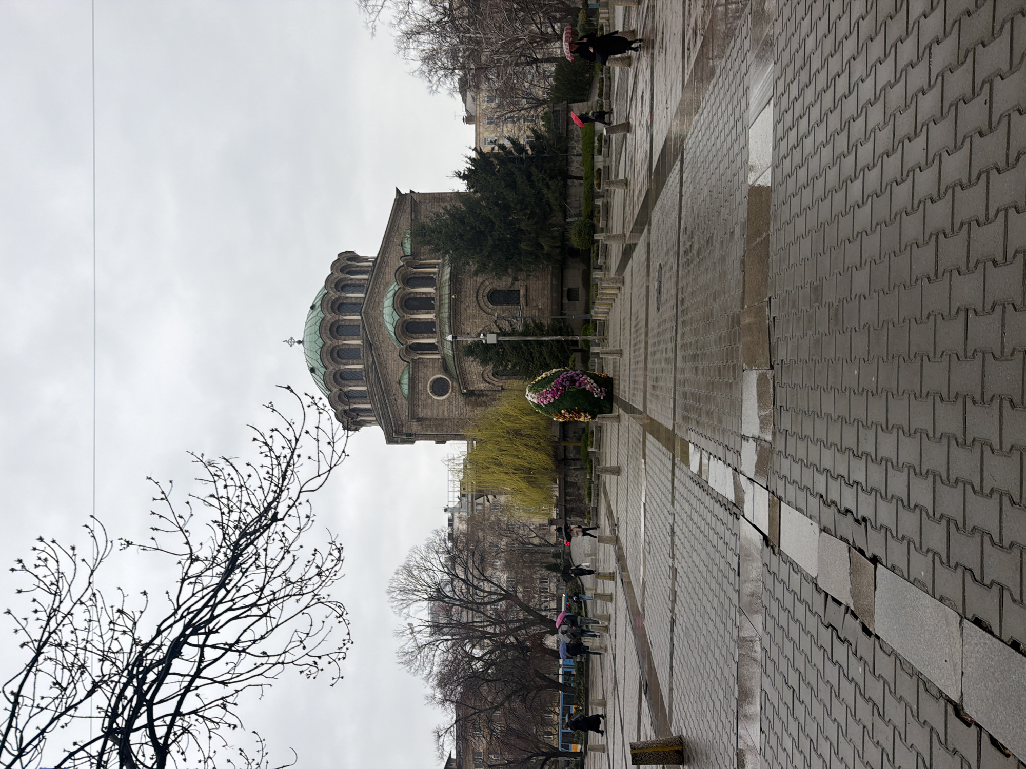 The image depicts a rain-slicked public square in Sofia, Bulgaria, dominated by the Alexander Nevsky Cathedral. The cathedral, constructed of stone, exhibits prominent domes and arched windows. Pedestrians holding umbrellas, some of which are colored, are visible traversing the square. A leafless tree with bare branches is present in the foreground. The ground is paved with interlocking concrete pavers, with areas showing water reflection. A flower bed containing a variety of colorful flowers sits at the end of the path. The sky is overcast, contributing to the dim lighting of the scene.