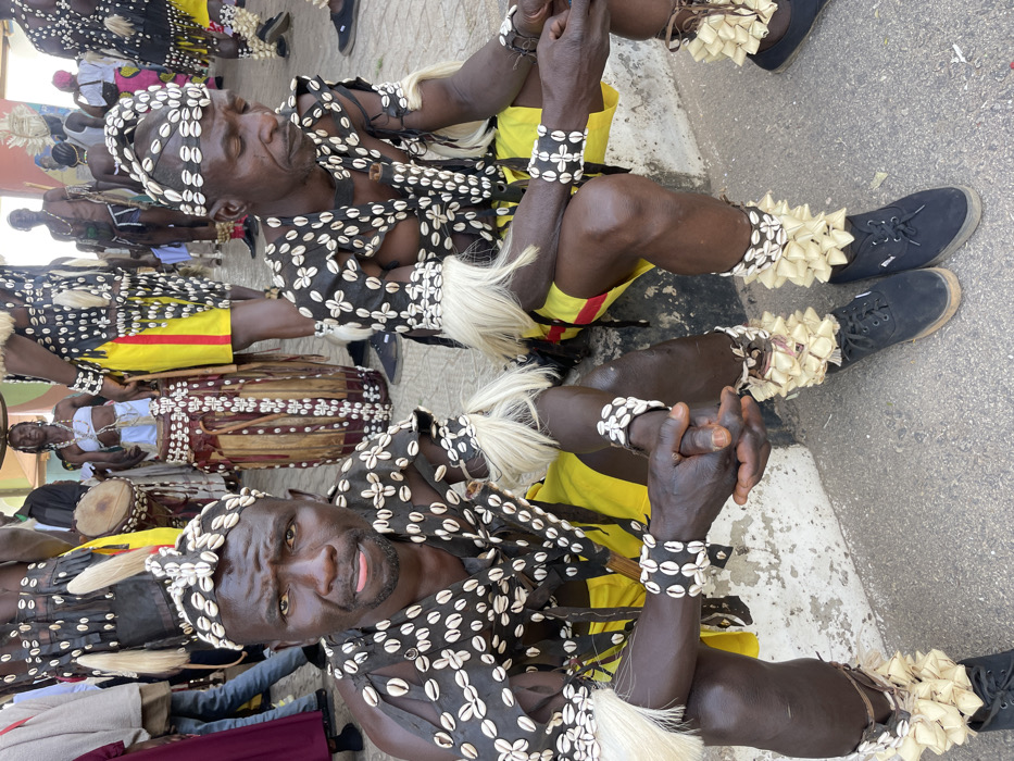 This image depicts a cultural gathering in Jos, Nigeria, showing two men in prominent traditional attire seated on a low concrete curb. The man on the left faces the viewer with a neutral expression, wearing a cowrie-shell-decorated headband, a black sleeveless top adorned with cowrie shells, yellow shorts, and white fur/feather armbands and ankle adornments over black lace-up canvas shoes. His hands are clasped. The man on the right, similarly dressed with a cowrie-shell headband, black and shell-decorated top, yellow shorts with a red stripe, white fur/feather armbands, and shell-decorated ankle wraps over black lace-up canvas shoes, looks down and slightly to his left, with his hands resting in his lap. The ground is a gray paved surface. In the background, other individuals are visible in various stages of similar traditional dress, including one person in the mid-ground playing a large, elaborately decorated drum with cowrie shells. The setting is an outdoor, public area.