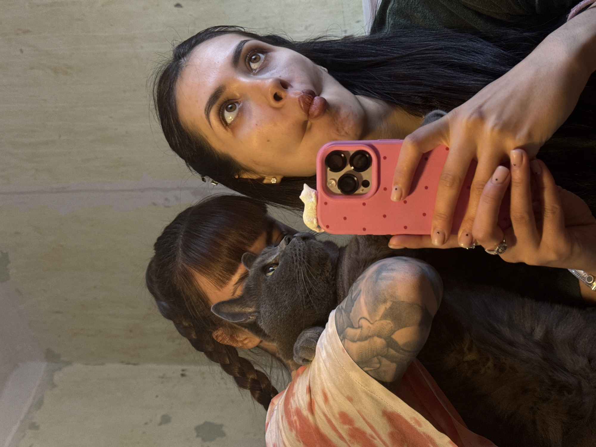 A woman is holding a pink cellphone with a white cat-shaped attachment and taking a selfie. She is pouting and looking upward. A gray cat is being held by another person in the frame. The other person has dark hair in a braid and a large tattoo on their arm. The background is blurry and appears to be a partially painted wall. The image does not contain sufficient information to ascertain the location in Votkinsk, Russia.