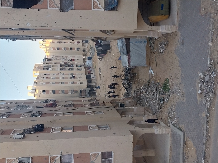 The image depicts a damaged urban residential area in Khan Yunis, Palestinian Territory, viewed from an elevated position between two multi-story, light-colored apartment buildings. These buildings exhibit visible structural damage, including shattered windows, exposed interiors, and missing sections, with rebar protruding in some areas. Clothes are seen hanging on balconies and from windows.
The ground between the buildings is an open, sandy, and debris-strewn space. In the foreground, a paved sidewalk and street surface are littered with small rocks and concrete fragments. Under the ground floor arches of the left building, a lone individual in dark clothing walks away from the camera. Under the corresponding arches of the right building, a vehicle covered with a yellow-green tarp is parked.
In the central dirt area, a charred wreckage, possibly a vehicle, lies amidst piles of rubble and debris. To its right, a makeshift shelter constructed from tarpaulins, one of which clearly displays a partial UNICEF logo, is visible. Further into the middle ground, a group of approximately ten individuals, mostly men in dark clothing, are gathered in a cluster on the dusty ground, appearing to observe or interact with the immediate surroundings. Additional figures are visible in the background, near other similarly damaged buildings that line the street beyond. The sky above is clear.