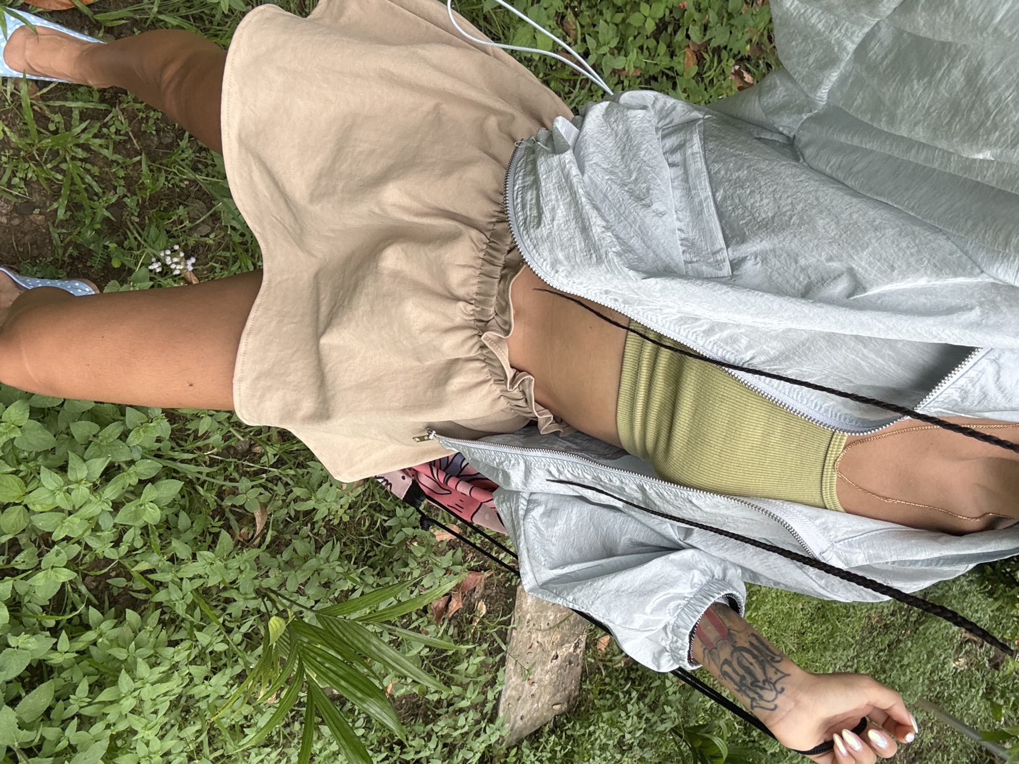 A person stands outdoors in a grassy area. The person is wearing a light blue windbreaker jacket over a lime green tank top and a beige skirt. The person has dark braided hair and tattoos on their left arm. Their feet are adorned with light blue polka-dot shoes. The background consists of lush green vegetation, including grass, plants, and a fallen log. The image appears to be taken from a slightly elevated, downward-facing angle, focusing on the person's midsection and legs. The overall setting suggests a casual outdoor environment, possibly a garden or park within Cẩm Thạch Commune, Vietnam.