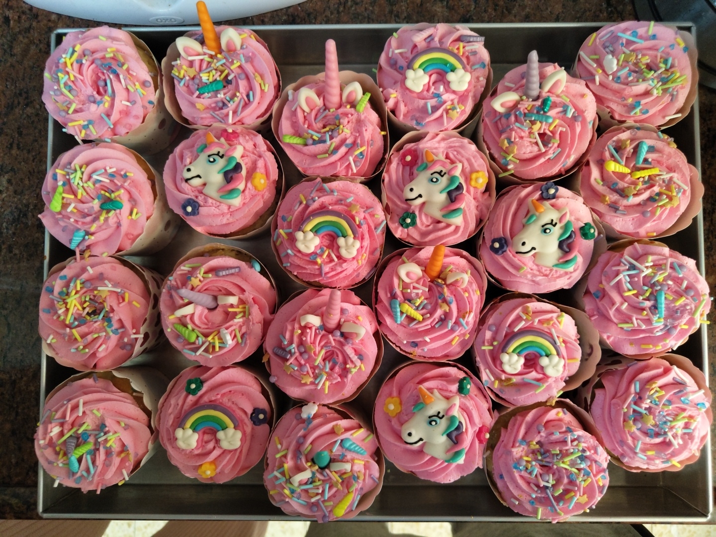 A metal tray contains 24 cupcakes. The cupcakes are decorated with pink frosting and colorful sprinkles. Some cupcakes have unicorn decorations. Others have rainbows and unicorn horns. The location is not identifiable from the image.
