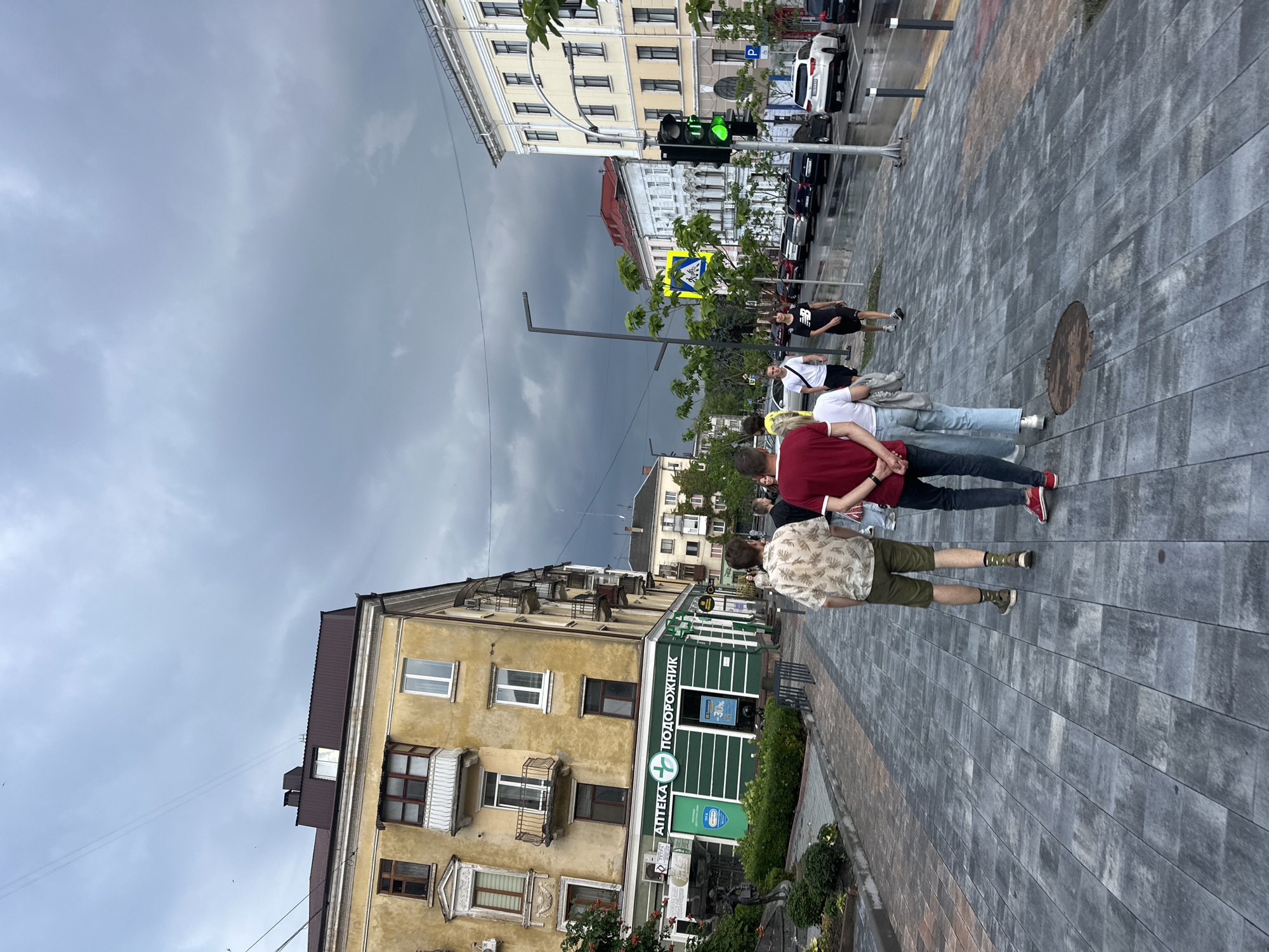 The image captures a street scene in Ternopil, Ukraine, under a dramatic, dark, and cloudy sky, suggesting impending or recent rain. The foreground is dominated by a wide pedestrian walkway paved with grey interlocking rectangular stones, some of which appear wet. Several individuals are walking away from the camera on this walkway.
To the left, a yellow building with multiple stories features balconies and windows. Prominently displayed on the ground floor is a green storefront with white lettering that reads "АПТЕКА ПОДОРОЖНИК," indicating a pharmacy. This building is adjacent to a small green area with bushes and a statue.
To the right, across the street, a light-colored building with multiple windows and an ornate facade stands. A traffic light shows a green signal. Cars are parked along the street, and the road surface also appears wet. Other buildings of varying architectural styles and colors are visible further down the street, creating a typical urban landscape. The overall atmosphere is somewhat somber due to the dark clouds, contrasting with the activity of people on the street.