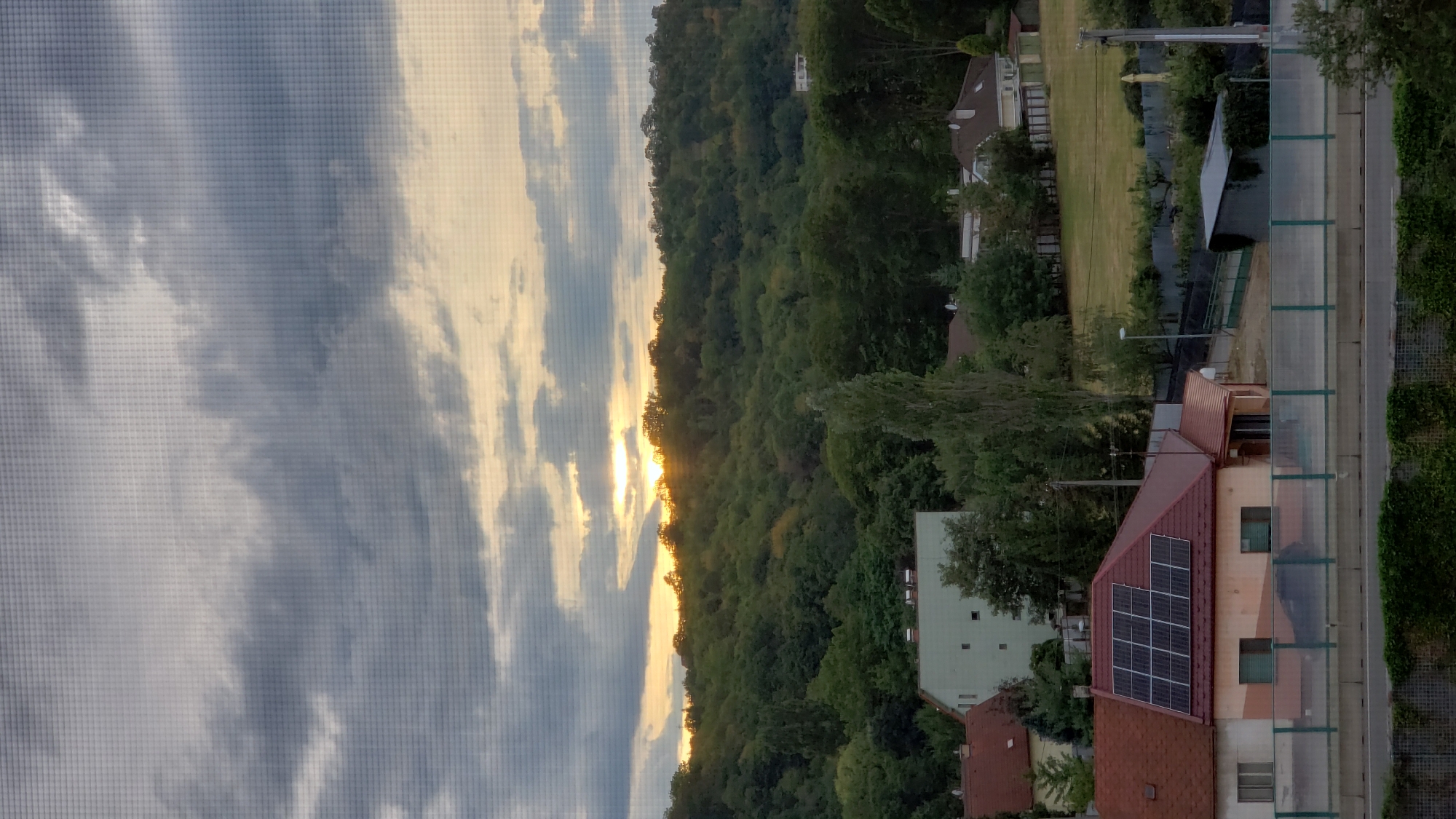 The image captures a sunset over a densely wooded hill, viewed through a window screen, which creates a grid pattern across the entire scene. In the foreground, a residential area is visible, featuring houses with red roofs. One house in the mid-foreground is particularly distinct, displaying solar panels on its roof. A highway barrier runs horizontally across the lower portion of the image, separating the residential area from a road. The sky is partly cloudy with the sun setting behind the trees on the hill, casting a warm glow. The surrounding environment appears to be a suburban or semi-rural area.