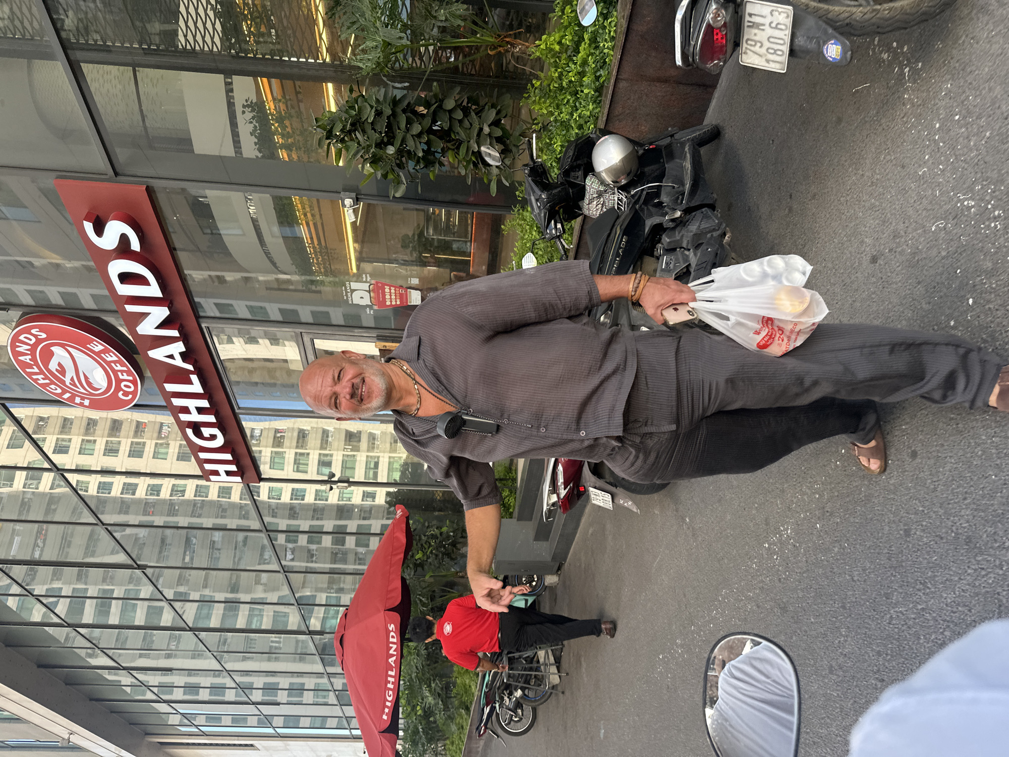 A bald, middle-aged Caucasian man is standing on a paved street in front of a modern building with a glass facade. He is wearing a grey, semi-sheer, button-down shirt and matching grey pants. He has a beaded necklace and several bracelets on his left wrist. In his right hand, he is holding a clear plastic bag containing two round objects, one orange and one white, and a smaller white item. His left arm is extended outwards, with his hand pointing towards the left.
Behind him, a red umbrella with "HIGHLANDS COFFEE" printed on it is open, and a person in a red shirt and black pants is walking away from the camera. There are several motorcycles parked on the street. The building behind the man has a large sign that reads "HIGHLANDS COFFEE". The overall environment suggests an urban setting, possibly in Bac Nha Trang Ward, Vietnam, given the context of "Highlands Coffee," a Vietnamese coffee chain.