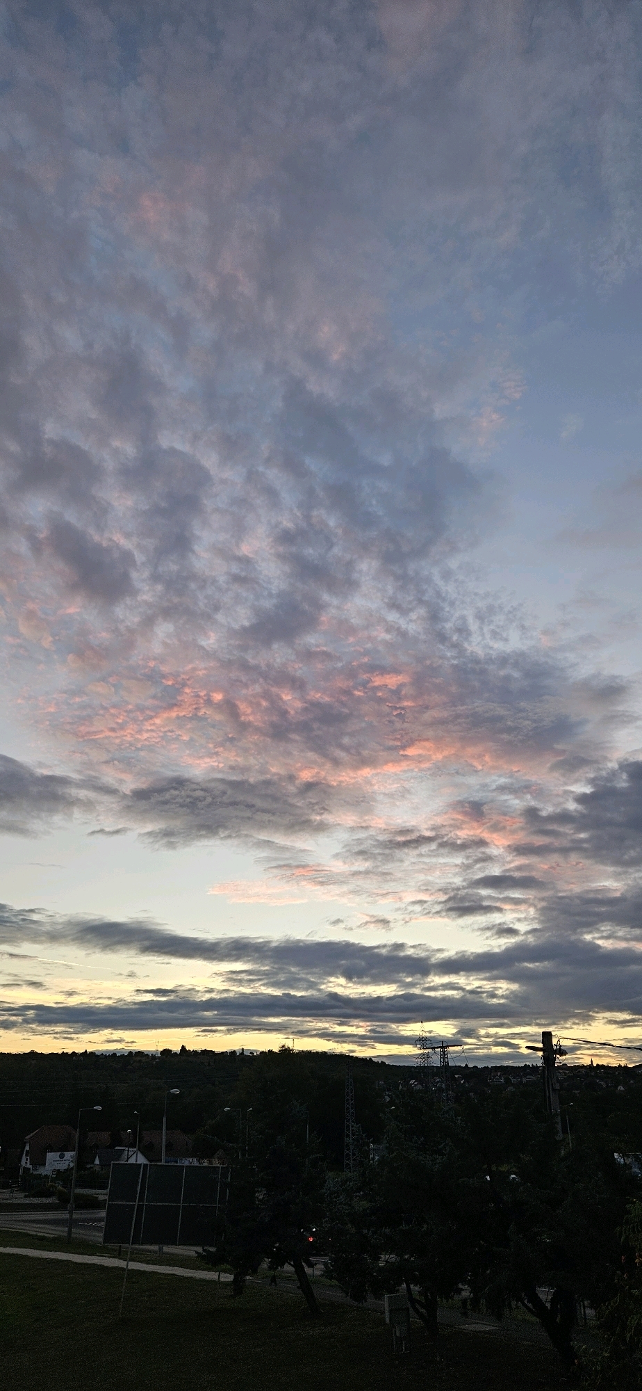 An outdoor, vertical, full shot captures a dusky sky over the Hungarian town of Alsópáhok. The sky is filled with wispy clouds, tinged with soft pink and orange hues from the setting sun, contrasting with the deeper blues and grays of the upper atmosphere. Below the sky, a dark silhouette of trees and hills forms the horizon line.
In the mid-ground, the outlines of buildings and structures are faintly visible, partially obscured by the trees. A few streetlights are visible, casting a dim glow. The foreground is dominated by dark trees and a patch of grassy ground, suggesting a park or natural area within the town. The overall impression is one of a quiet evening, with the natural beauty of the sky taking center stage.