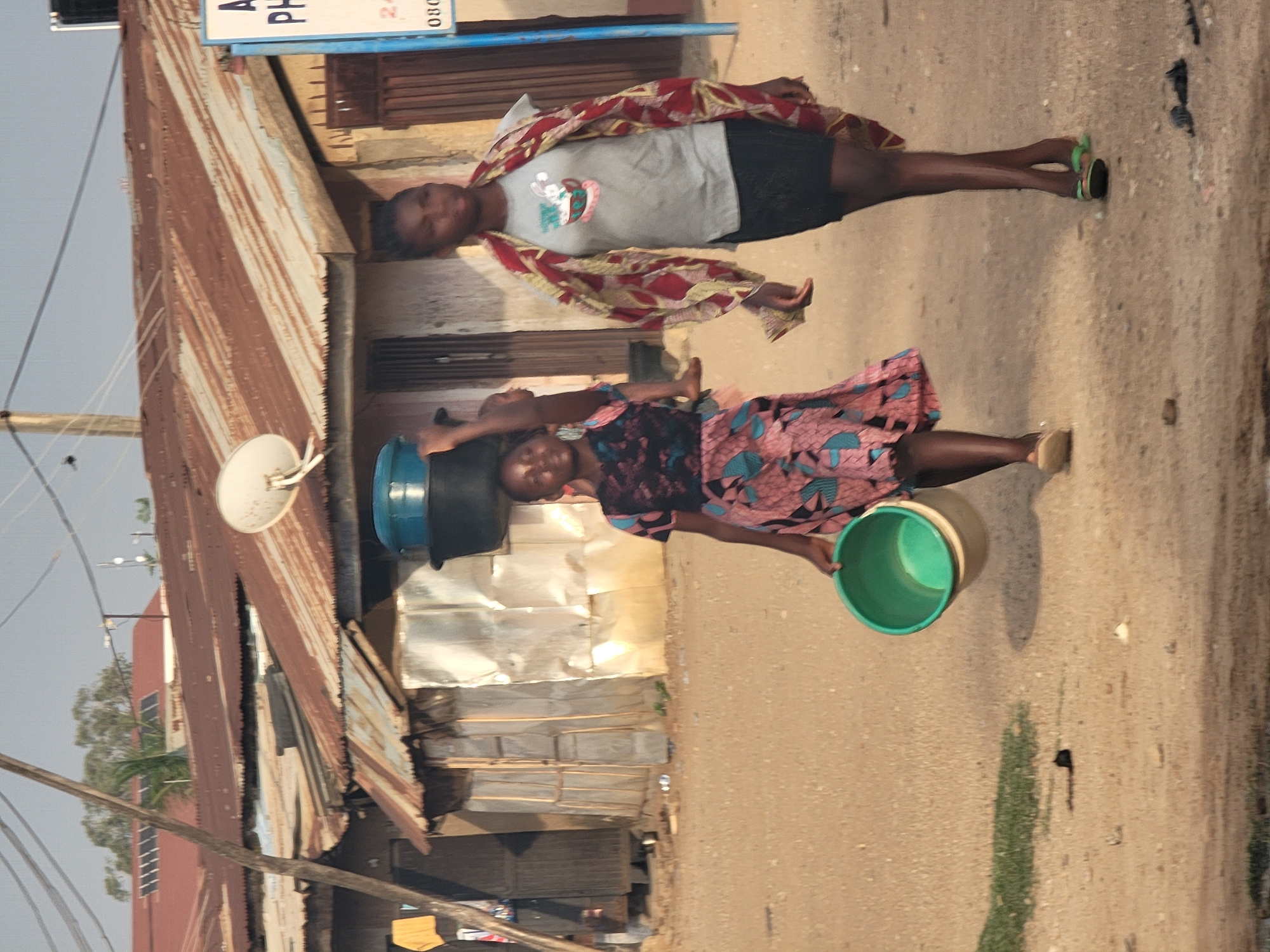 Two young women are walking on a dirt road in Sabon Tasha, Nigeria. The woman closest to the viewer is carrying a green bucket in her right hand and a blue and black bucket on her head. She wears a patterned dress and sandals. The other woman is wearing a gray t-shirt, black shorts, a patterned shawl, and green sandals. Both women are walking away from the camera. A dilapidated building made of corrugated metal is on the left side of the road, with a satellite dish attached to it. There are power lines and utility poles in the background. Vegetation and other buildings are visible in the distance.
