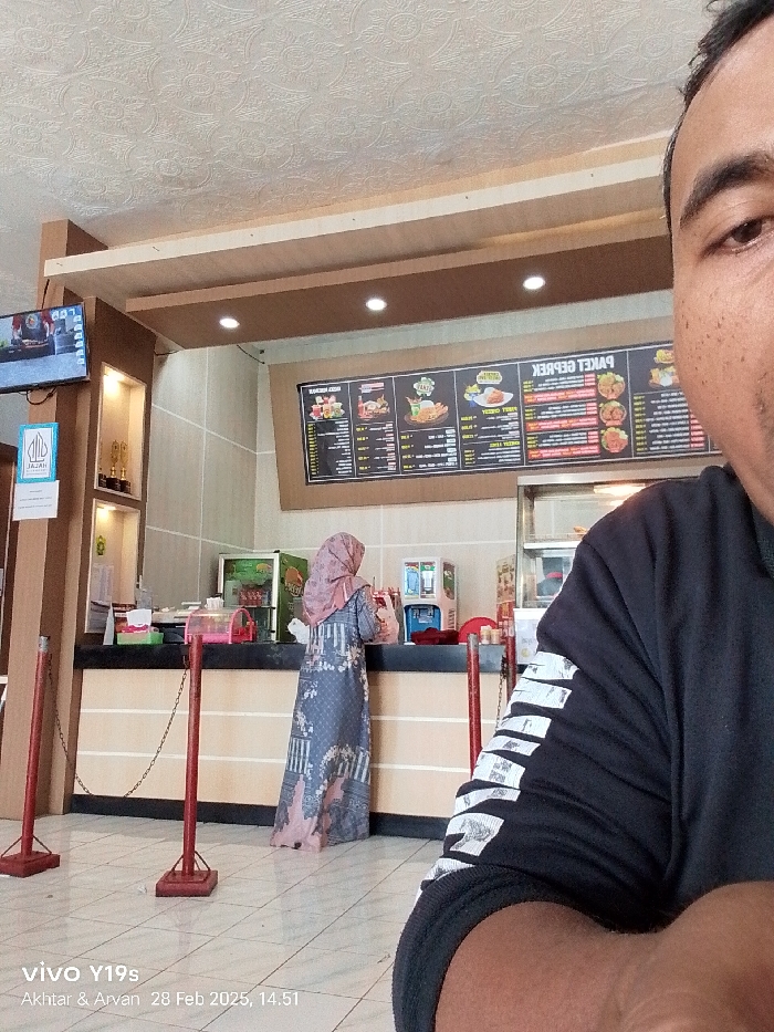The image depicts a scene inside a restaurant located at latitude -6.318 and longitude 107.986 in the city of Patrol, Indonesia. A woman in a long floral dress is standing at a counter. She is facing away from the camera. The counter is topped with a large metal chain which is attached to red poles on either side. Behind the counter, a menu board lists various food items. The menu is written in Indonesian. An individual wearing a black shirt with a white stripe design is partially visible in the right foreground, covering most of the camera's view. The individual's hand is resting on the counter. The environment is well-lit with fluorescent ceiling lights. The floor is tiled with square gray and beige tiles. The walls are light beige and have a textured design. A large television monitor is visible on the wall to the left of the counter. It is displaying a black and white video. A sign in the foreground displays a graphic image of a hand and the word "Jaga". The image appears to be taken from a cell phone camera, and the text on the bottom of the photo indicates the date and time as 28 Feb 2023, 14.51. There are other people in the restaurant but they are not visible in the picture.