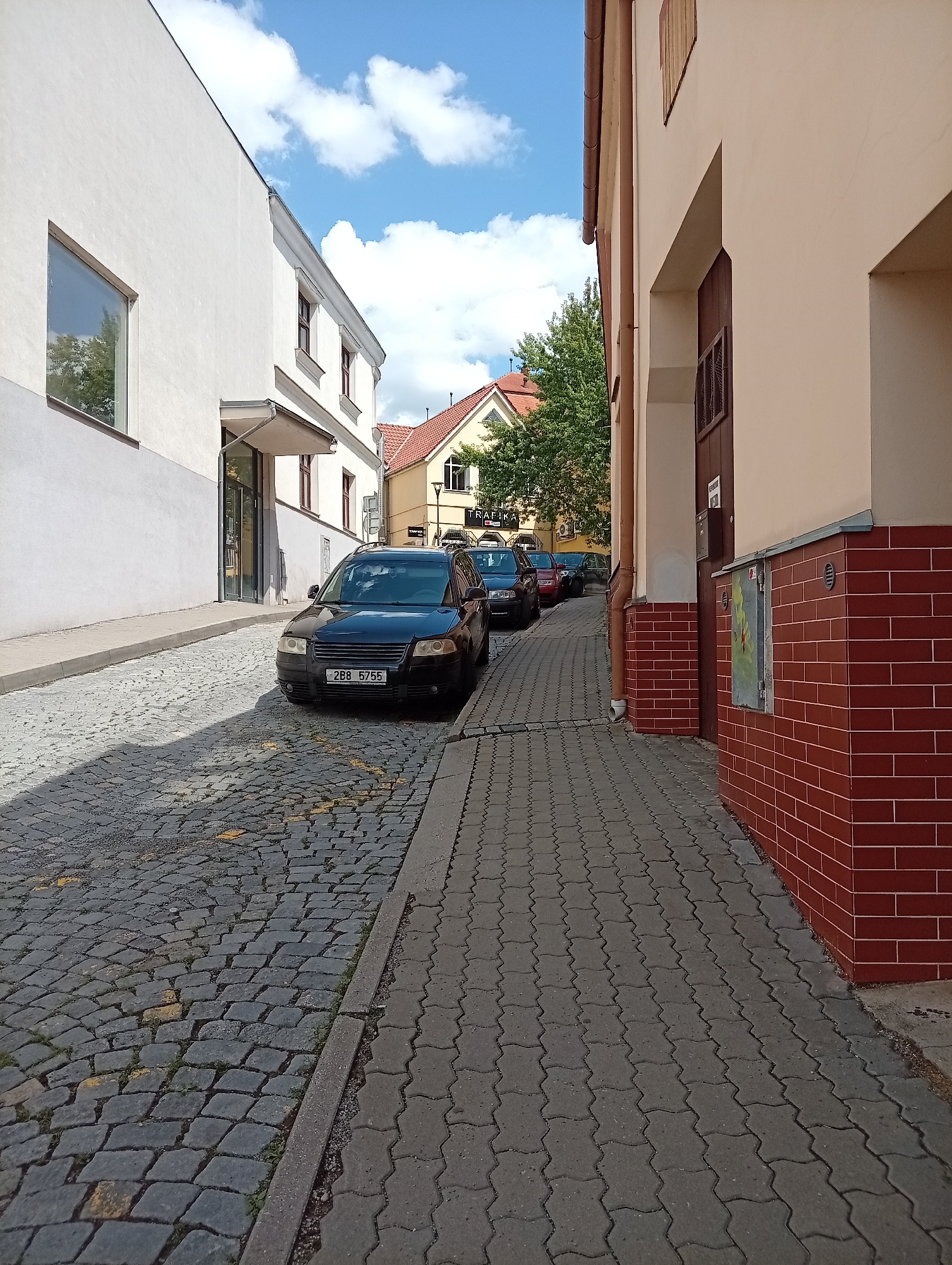 The image depicts a street in the city of Tišnov, Czechia, on a sunny day with a blue sky and scattered clouds. The street is paved with cobblestones on the left side and interlocking concrete pavers on the right side. A row of parked cars is visible on the right side of the street. The buildings on either side of the street are typical of a European town, with one building being a light-colored, modern structure with a large window, and the other being an older building with a beige facade and red brick detailing. There are trees and glimpses of other buildings in the background, suggesting a residential or commercial area. The overall impression is of a quiet, sun-drenched street in a European town.