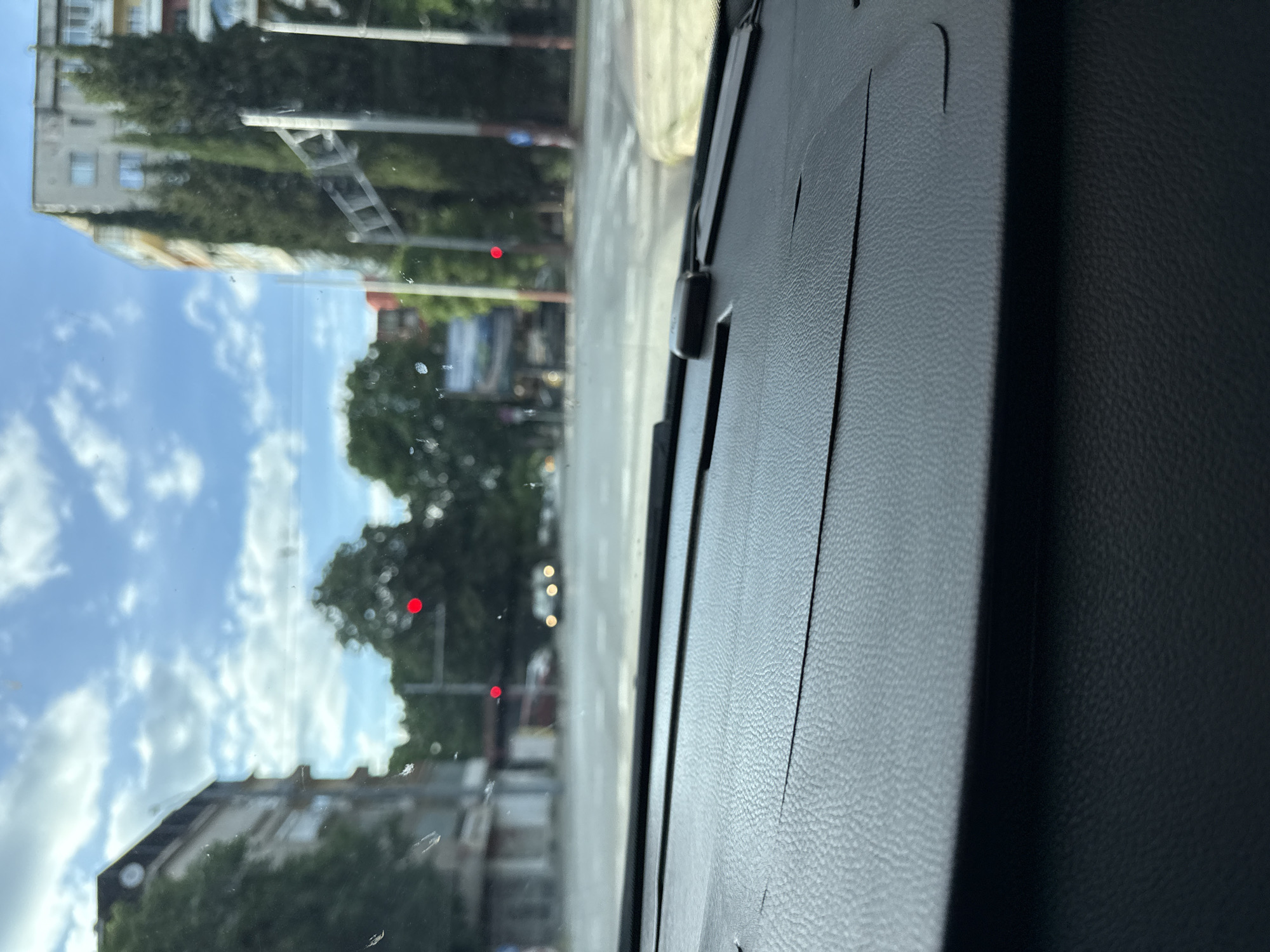 The image shows a view from inside a vehicle, looking out onto a street scene in Stara Zagora, Bulgaria. The dashboard of the vehicle is visible in the foreground. Through the windshield, the image shows a street with buildings on either side, trees, and a traffic light showing red. The sky is blue with some clouds. There are no visible people. The environment is an urban setting during daylight hours.