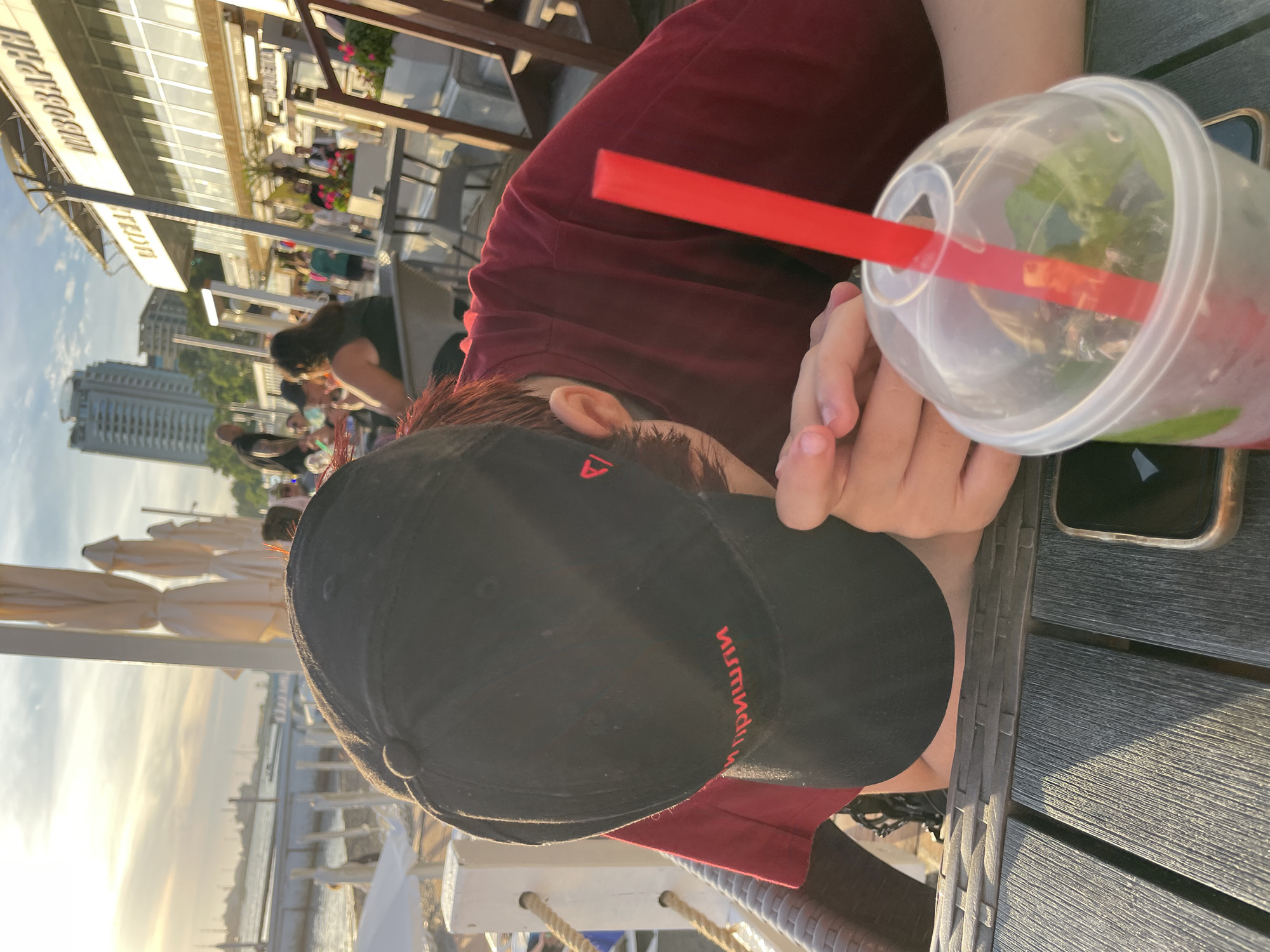 The image shows a close-up of a person wearing a black baseball cap and a maroon t-shirt, seated at a table outdoors. The person's face is mostly obscured by the cap, but their ear is visible. A clear plastic cup with a red straw and a green beverage is placed on the table, along with a smartphone. The table appears to be made of dark wood. In the background, there are glimpses of other people, umbrellas, and buildings, suggesting an outdoor seating area of a cafe or restaurant. The lighting indicates it is either early morning or late afternoon, with a warm glow on the scene. The overall setting suggests a relaxed, casual atmosphere. There is text visible on the brim of the cap, in red lettering, which appears to be in Cyrillic. There is also some text on a sign in the background, which is partially visible and also appears to be in Cyrillic. The scene is likely in a coastal area, given the presence of water and boats in the distance.