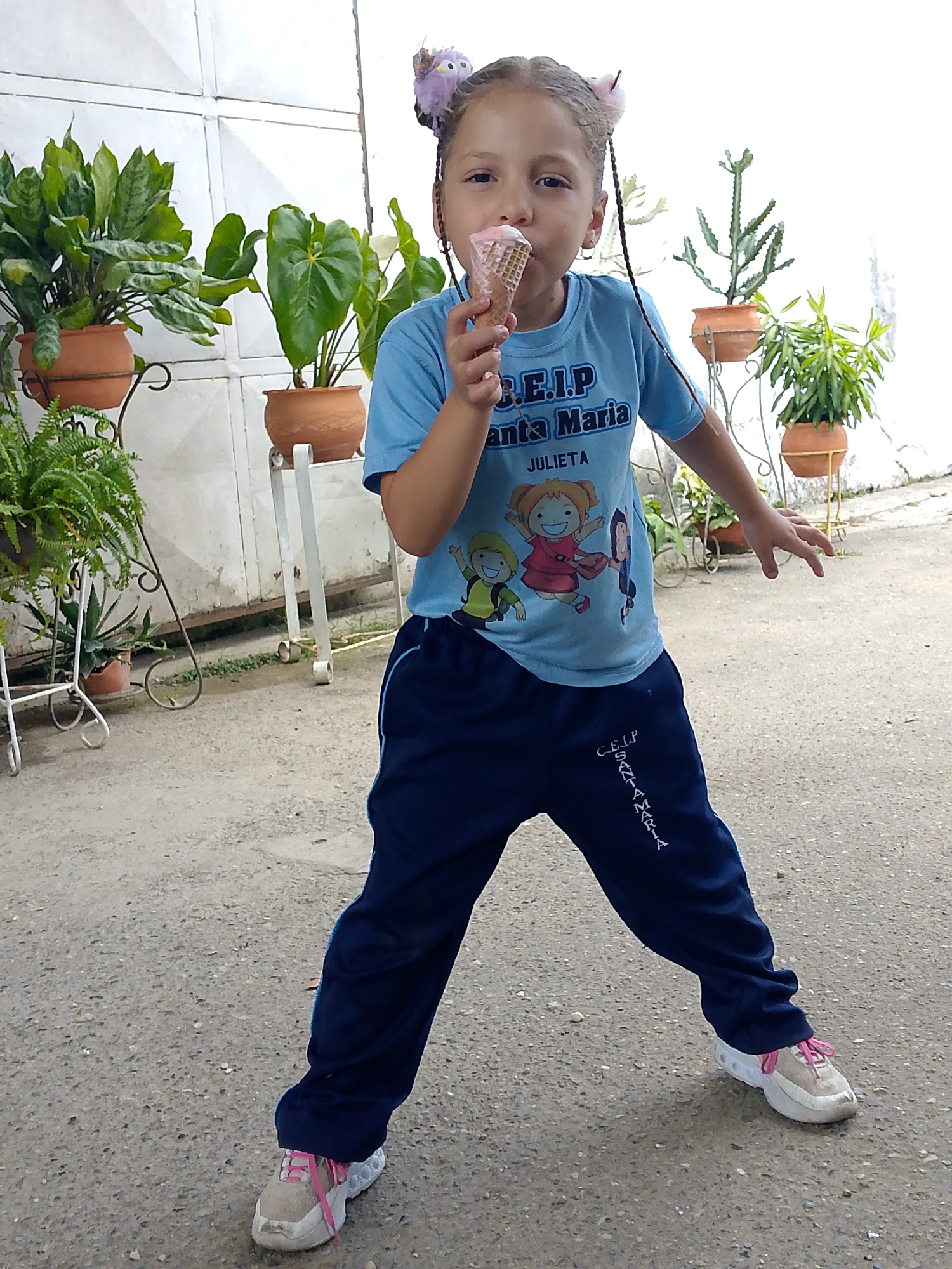 A young girl is standing outdoors, eating an ice cream cone. She is wearing a light blue short-sleeved shirt with a print of cartoon figures and the words "C.E.I.P. Santa Maria Julieta," and dark blue sweatpants with the words "C.E.I.P. Santa Maria" printed down the leg. Her shoes are white with pink laces. The girl has braids and hair accessories. She is standing with her legs apart and one arm outstretched. In the background, there is a white wall, various potted plants on stands, and a concrete surface. The image was taken in Tinaquillo, Venezuela.