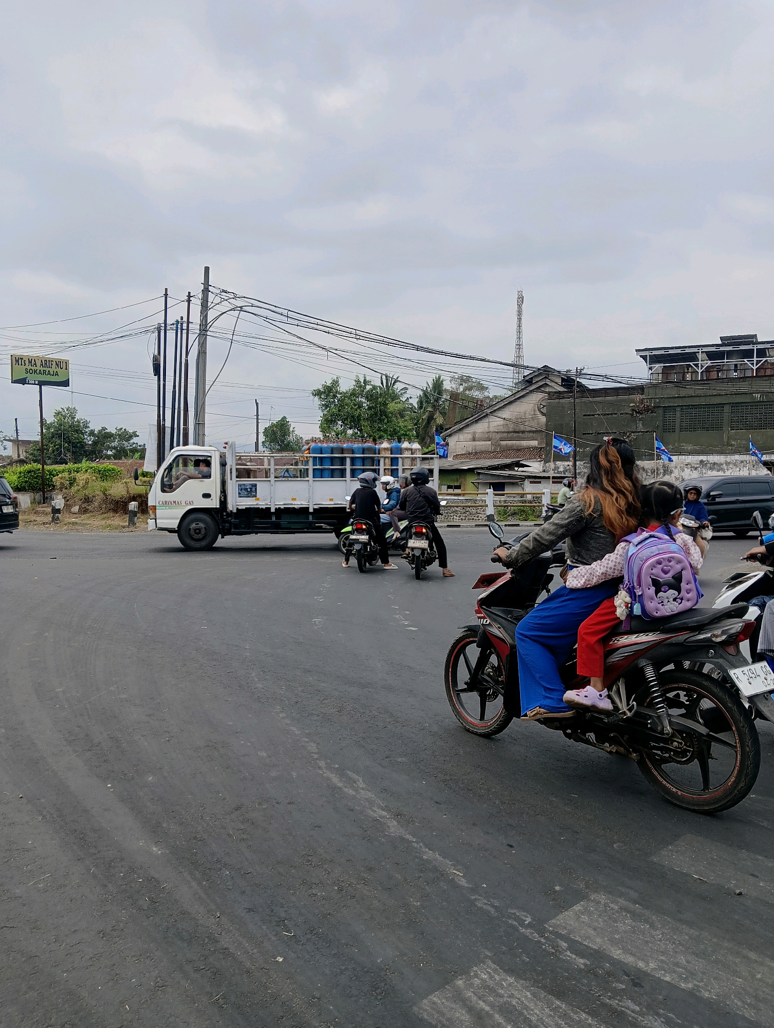 A street scene in Sokaraja, Indonesia is depicted under an overcast sky. In the foreground, a motorcycle with two passengers, a woman and a young girl, is in motion. The woman, wearing a gray jacket and blue pants, is operating the motorcycle, and the girl, in a red outfit and carrying a purple backpack with a cartoon cat design, is seated behind her. The motorcycle has red accents on its wheels. To the left, a white truck is driving, carrying numerous blue gas cylinders in its bed. The truck has "CARTAMAS GAS" written on its side. Several other motorcycles are also visible in the background, along with a black car. A green sign with white text reading "MTs MA ARIF NU 1 SOKARAJA" is positioned on the left side of the road, indicating the location. The environment includes a paved road, overhead power lines, buildings, trees, and streetlights. The road surface shows signs of wear and tire marks.