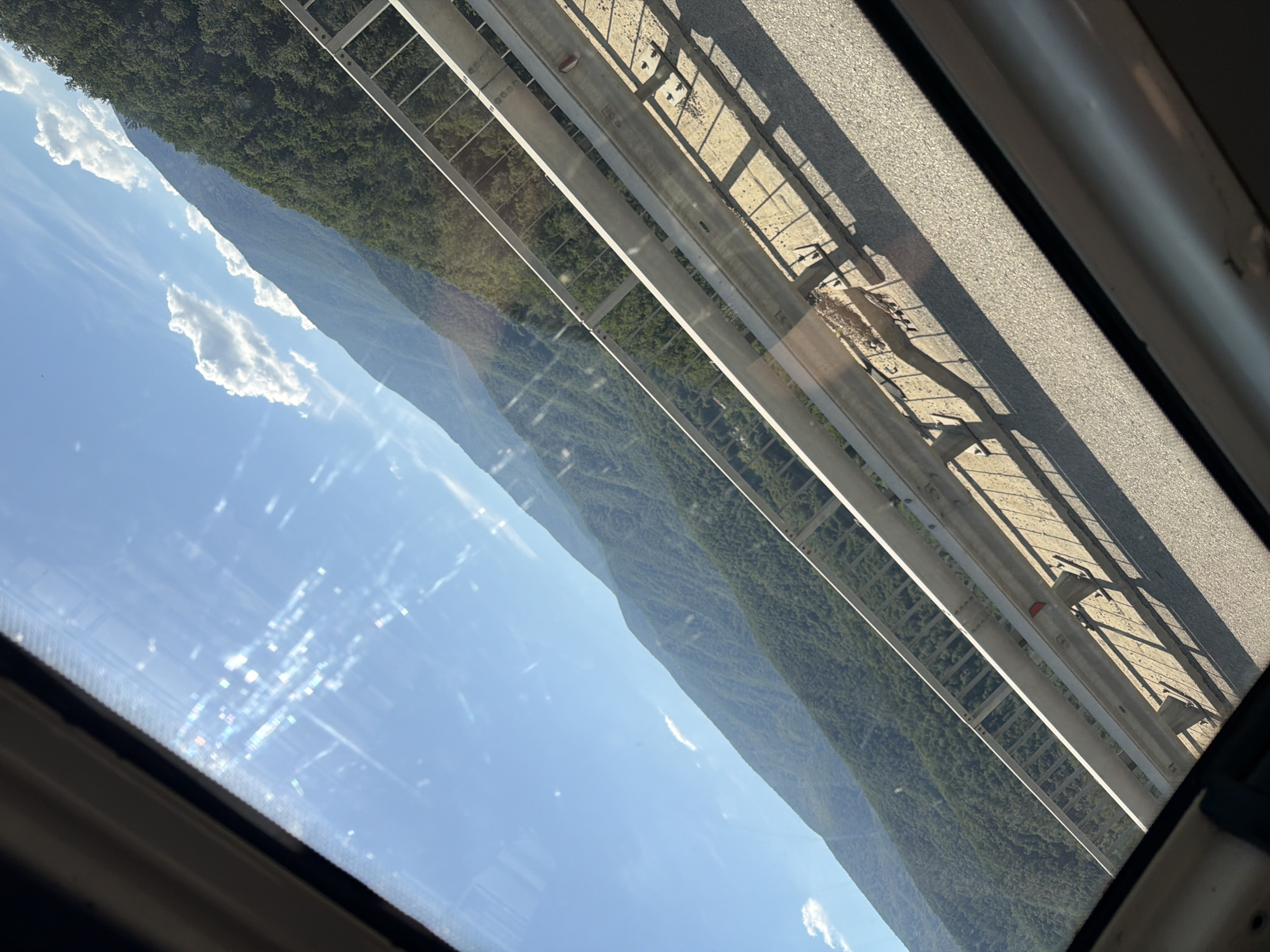 The image is a photograph taken from inside a vehicle, looking through a window. The view is angled, showing a mountainous landscape under a bright blue sky with scattered white clouds. In the foreground, a metal guardrail runs along the edge of what appears to be a road or bridge, casting shadows on the asphalt. The mountains are covered in dense green forest. The sun is shining brightly, causing lens flare and reflections on the window. The image appears to be taken on a sunny day in the vicinity of Vrachesh, Bulgaria.