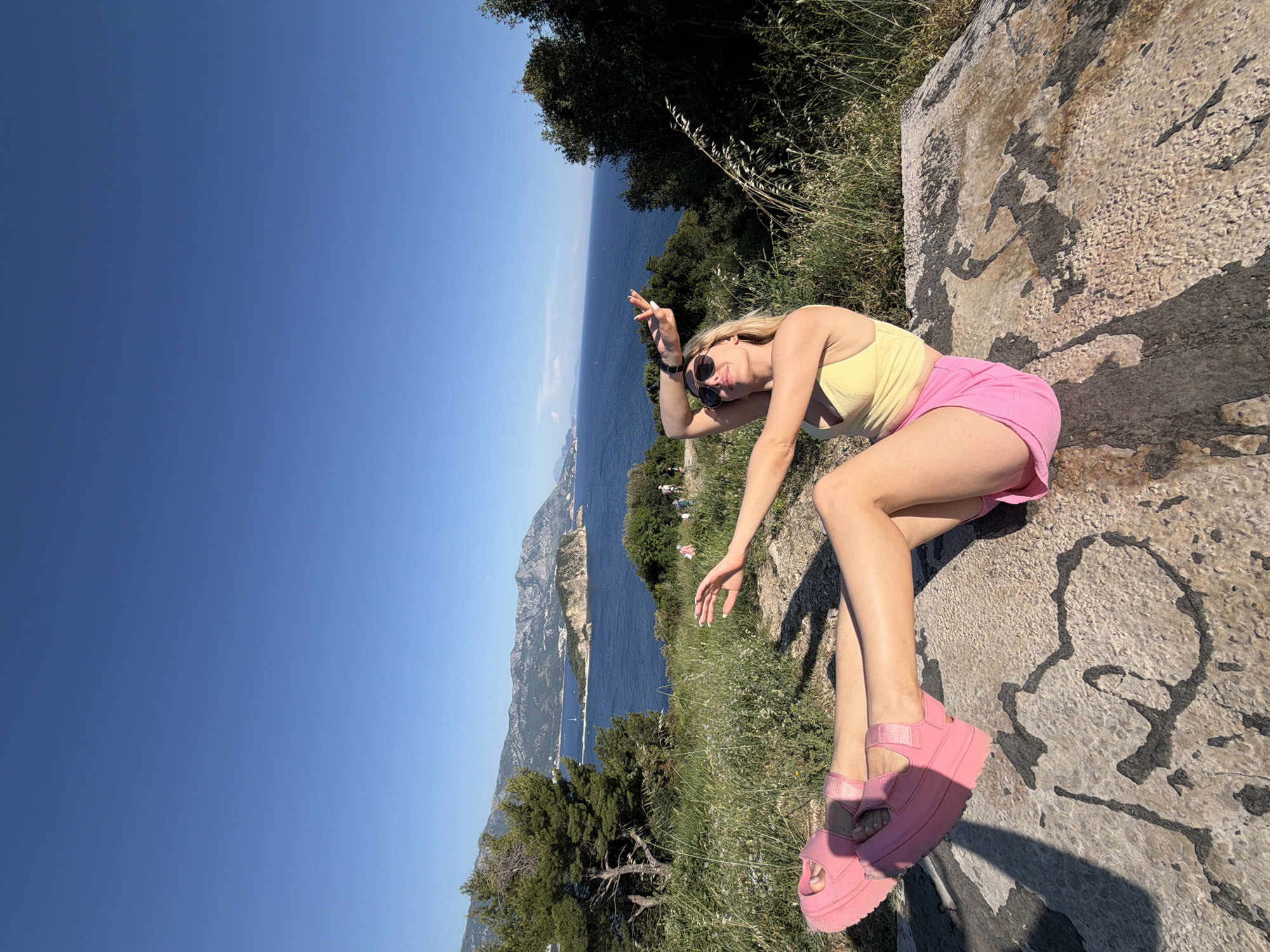 The image shows a woman reclining on a stone surface, likely a viewpoint or landmark. She is wearing sunglasses, a yellow top, pink shorts, and pink platform sandals. Her location overlooks a coastal landscape, with the sea, mountains, and some trees visible in the background. This setting is likely in the vicinity of the city of Budva, Montenegro, given the geographical context. The woman is posing with one arm raised and fingers extended. The sky is blue.