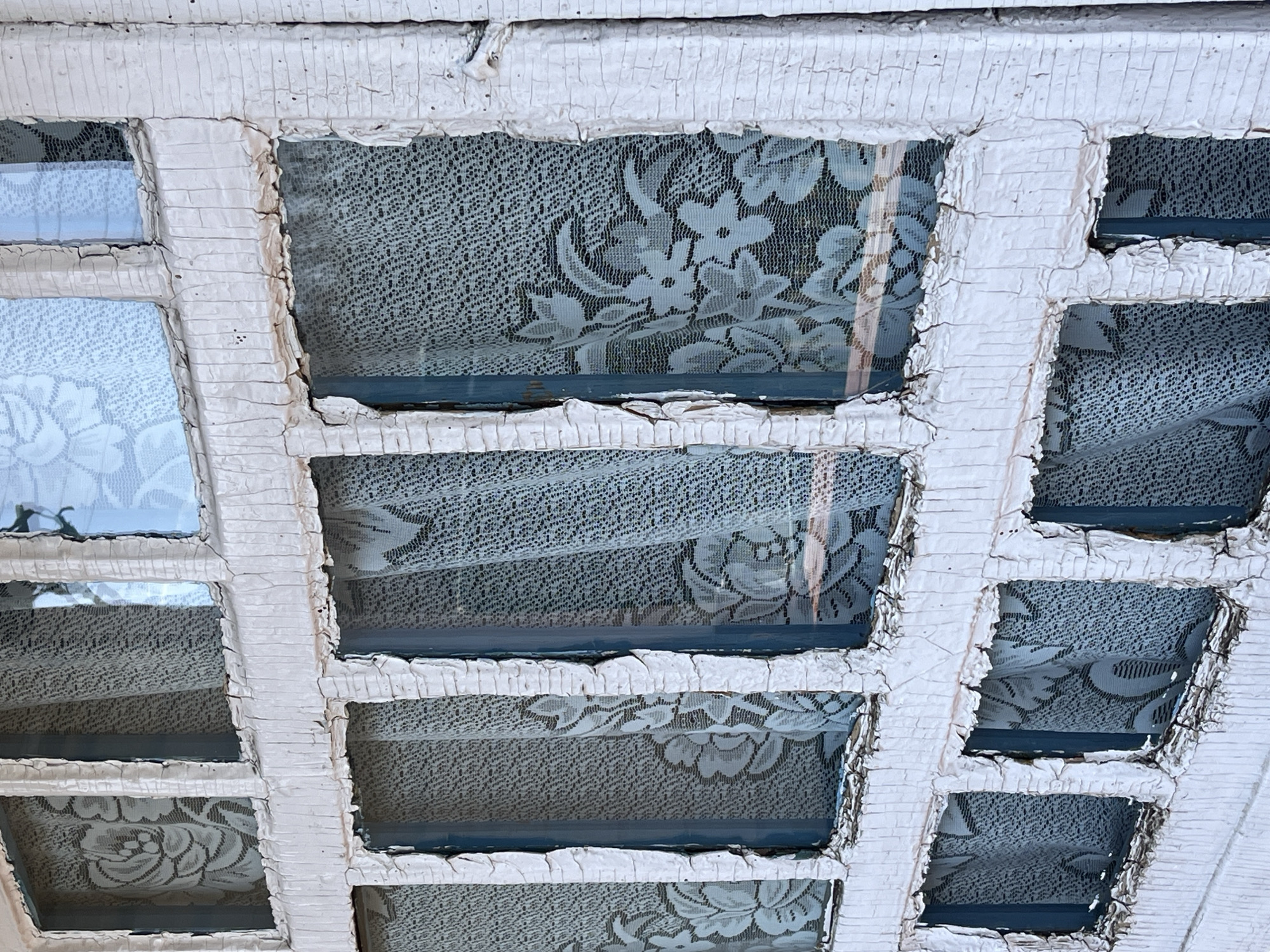 The image shows a grid of small, square windows with white, heavily cracked and peeling frames. The glass panes are partially obscured by lace curtains with a floral pattern. The environment is not directly visible beyond the curtains. The location is specified as Верхняя Уря, Russia, but there are no visual indicators in the image to confirm this.
