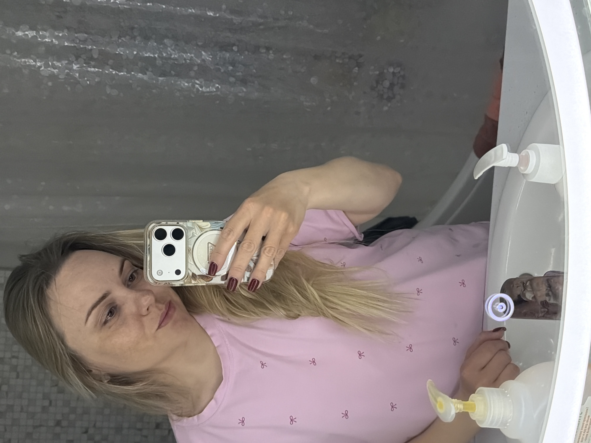 A woman is taking a selfie in a bathroom. The bathroom appears to be in a home. The woman is holding a cell phone in her right hand and is looking at the screen. She is wearing a pink shirt with small bows on it. The woman has blond hair that is partially draped over her shirt. The background includes a bathroom mirror, shower curtain, sink, and two bottles of soap. The location is Votkinsk, Russia, as inferred from the prompt.