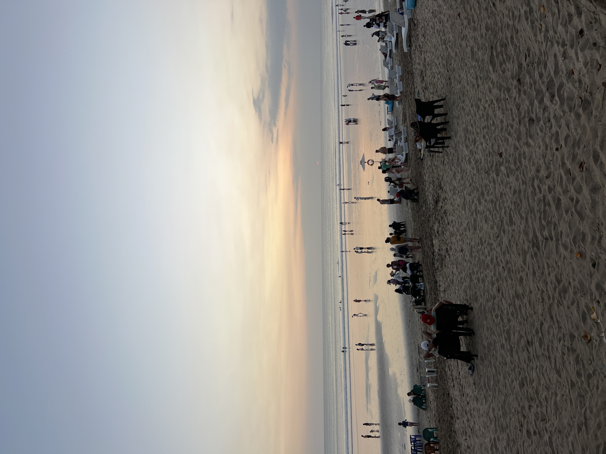 The image depicts a wide, sandy beach in Kuta, Indonesia, during a sunset. The sky is a gradient of soft blues, pinks, and oranges. The ocean is calm with gentle waves, and the wet sand reflects the colors of the sky, creating a mirror-like effect. Numerous people are visible on the beach, some wading in the shallow water, others walking along the shore, and groups gathered on the sand. There are lounge chairs and umbrellas set up in some areas, indicating a recreational setting. In the foreground, two individuals are seated on chairs facing away from the camera, overlooking the beach. The overall atmosphere is serene and leisurely, capturing a typical evening scene at a popular beach destination.