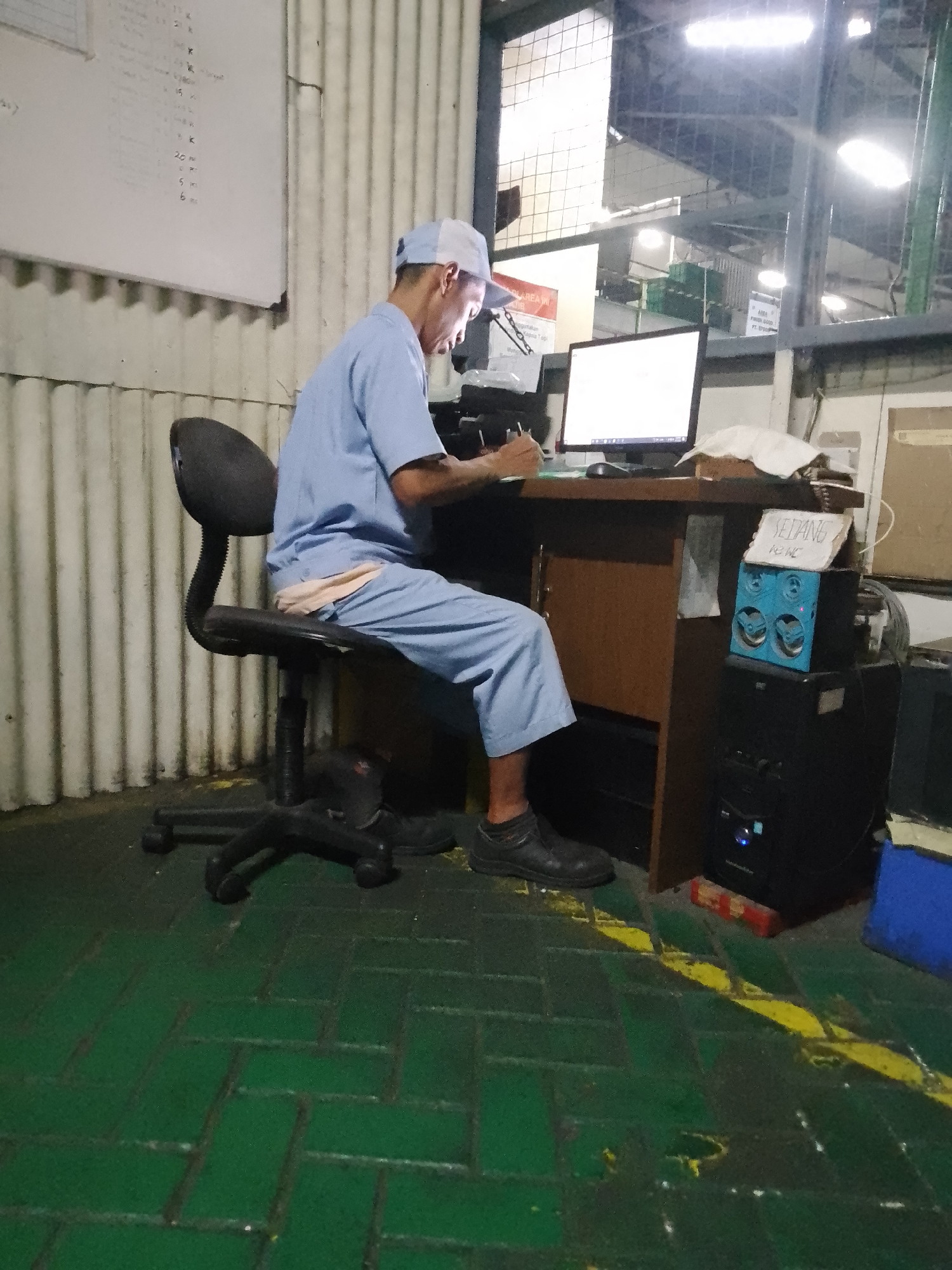 The image shows a person wearing a light blue uniform and a white and blue baseball cap, sitting at a desk in what appears to be a warehouse or factory setting. The person is looking down at a piece of paper and writing with a pen, with a computer monitor and keyboard in front of them. The desk is made of wood, and there is a black office chair.
To the right of the desk, there is a black computer tower with a blue speaker system on top. A small sign with handwritten text is placed on the speaker. The floor is green with yellow markings. In the background, a corrugated metal wall is visible, and a large window with metal bars looks out into another part of the facility, illuminated by bright artificial lights, suggesting it might be nighttime or an indoor environment with limited natural light. There's a whiteboard with some writing on the left side of the frame.
The overall scene suggests an individual working at a computer, possibly managing inventory or production data, in an industrial environment. The lighting indicates it could be late or an indoor work area.
Visible text: The sign on the speaker appears to read "SEDANG" and below it "RENC". The whiteboard on the left has handwritten notes, some of which look like numbers and letters. The computer monitor also displays text.