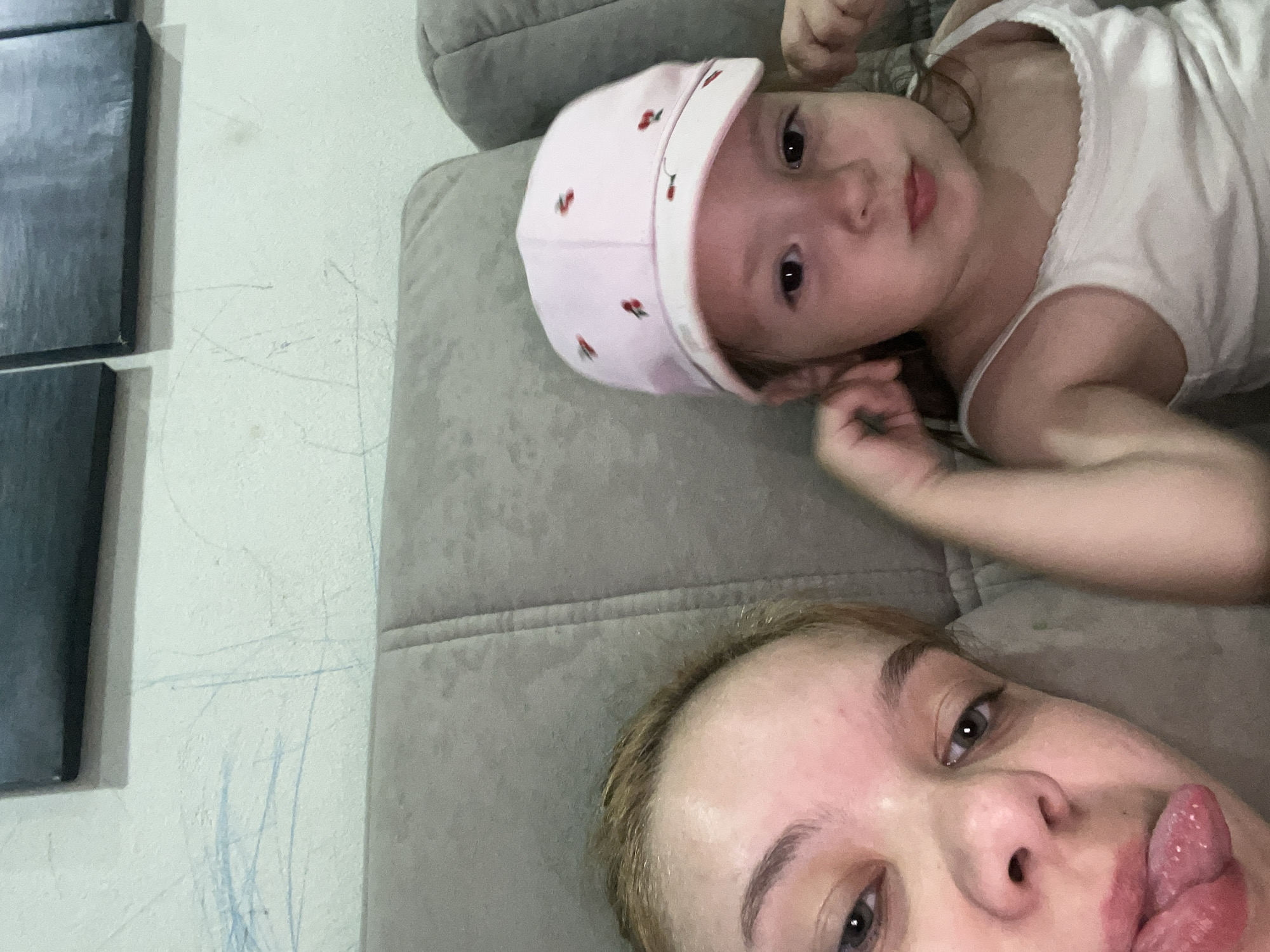 The image depicts two individuals, an adult and a young child, seated on a cushioned sofa. The adult, on the left, is closer to the camera and has blonde hair. The child, on the right, is wearing a pink cap adorned with small red fruit designs and a white tank top. The background shows a wall with some scuff marks, and above it, what appears to be a piece of dark artwork or a frame. The setting is indoors, likely a domestic environment. The image does not contain information about the city of Gorodets, Russia.