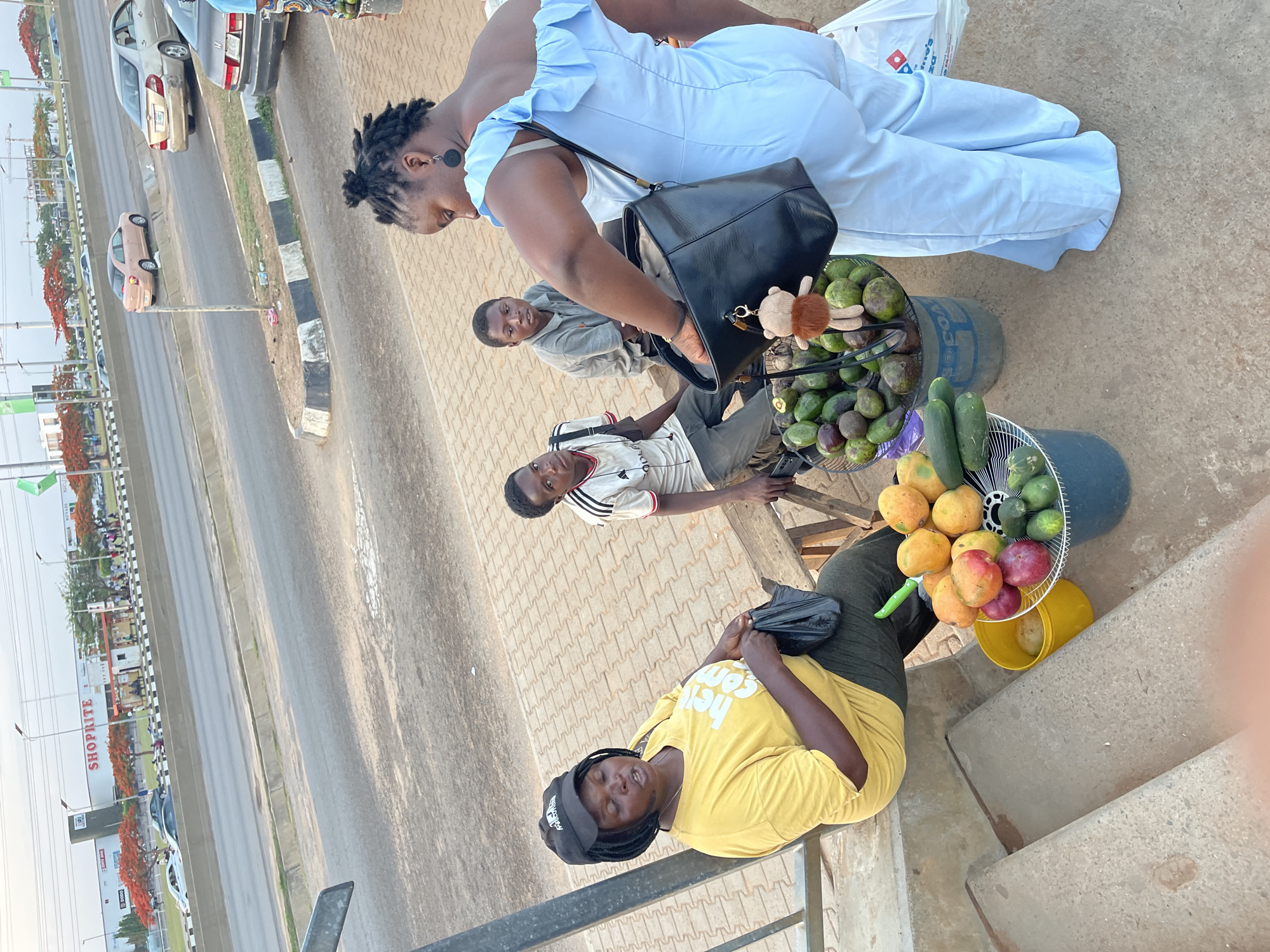 The image shows a street scene in Galadimawa Village, Nigeria. Three men and one woman are visible. The woman, wearing a light blue dress, is reaching into a black handbag. Next to her are two men sitting, and one is looking at the camera. In front of them are two baskets of fruit, including avocados and mangoes, sitting on top of buckets. At the bottom of the image, there is a woman wearing a yellow shirt and black baseball cap leaning against a railing. In the background is a street with cars driving, and a large Shoprite building.