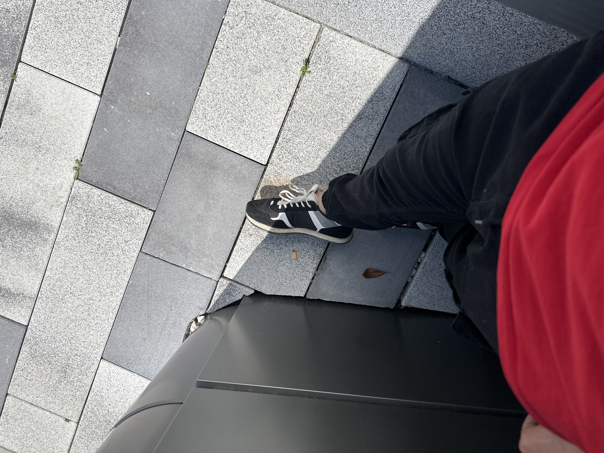 A high-angle shot depicts a person's legs and a shoe on a gray tiled surface. The person wears black pants and a red shirt. The left shoe is black with white stripes and laces, resting on the gray tiles that form a patterned walkway. Part of a black metallic structure is visible in the foreground. Based on visual patterns of the ground, the image is taken in the city of Lovech, Bulgaria.