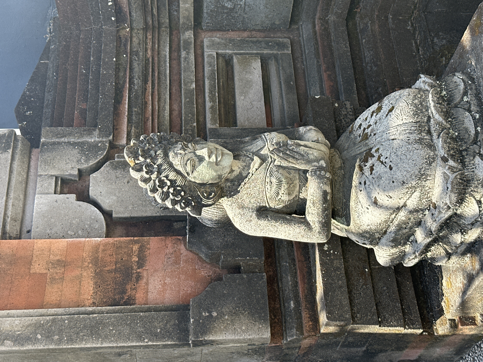 A stone statue of a woman in traditional Balinese attire is kneeling on a lotus-shaped pedestal. She has her hands pressed together in a prayer-like gesture. The statue is carved with intricate details, including a headdress adorned with flowers, a necklace, and a decorative bodice. The statue is situated in front of a stone wall with red brick accents. The wall is weathered and shows signs of age, with moss growing on some surfaces. The overall scene suggests a cultural or religious site, possibly in the Ubud District of Indonesia, known for its rich artistic and spiritual heritage.