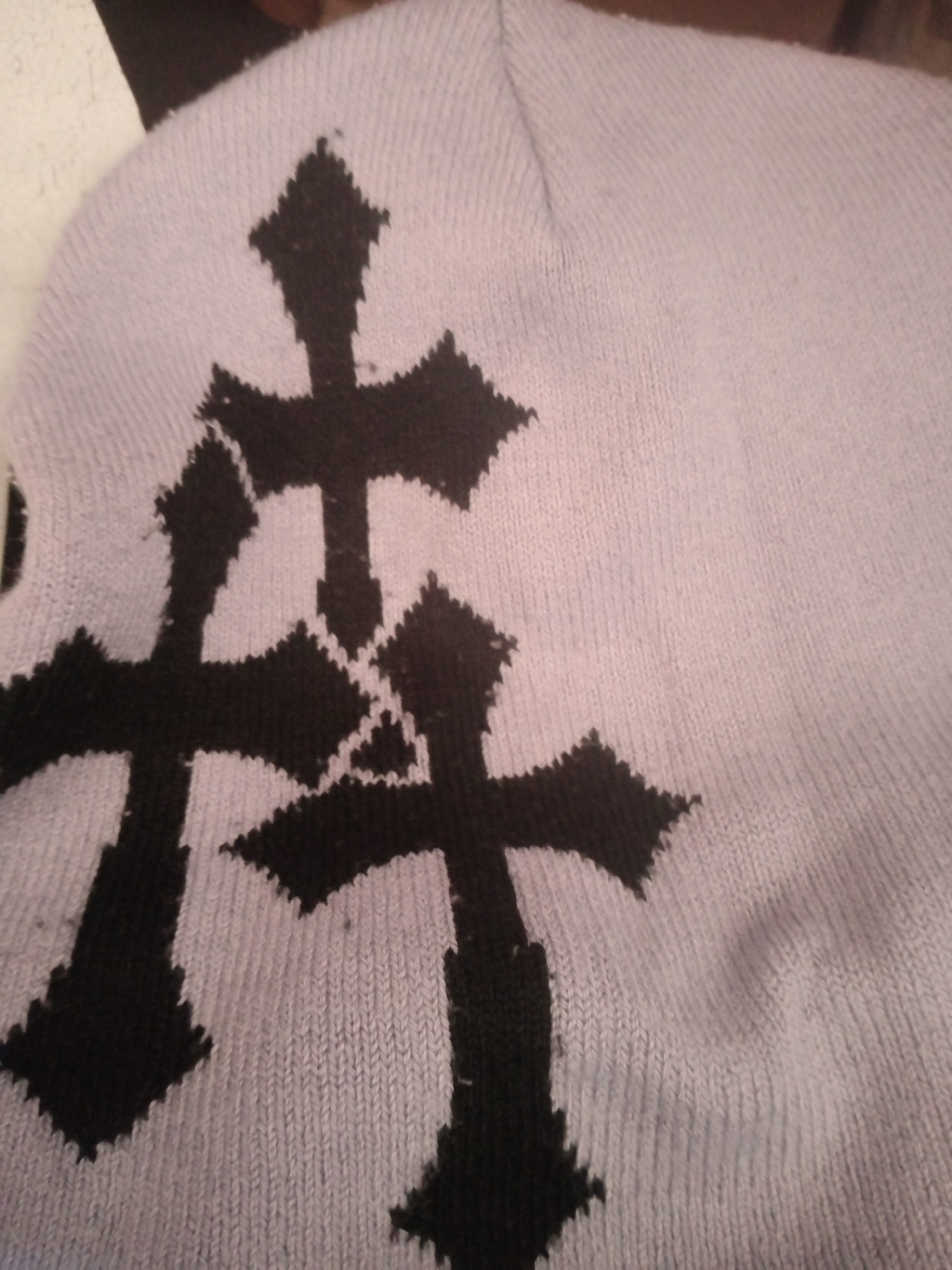 The image shows a close-up view of a light purple knitted fabric adorned with three black cross designs. One large cross is positioned centrally, with two smaller crosses partially visible below and to the left and right of it. The fabric appears to be part of a garment, possibly a sweater or a beanie, given its textured knit. The lighting is somewhat dim, creating soft shadows on the fabric's surface. There are no people, specific locations in Jalingo, Nigeria, or any discernible actions or interactions depicted in the image.