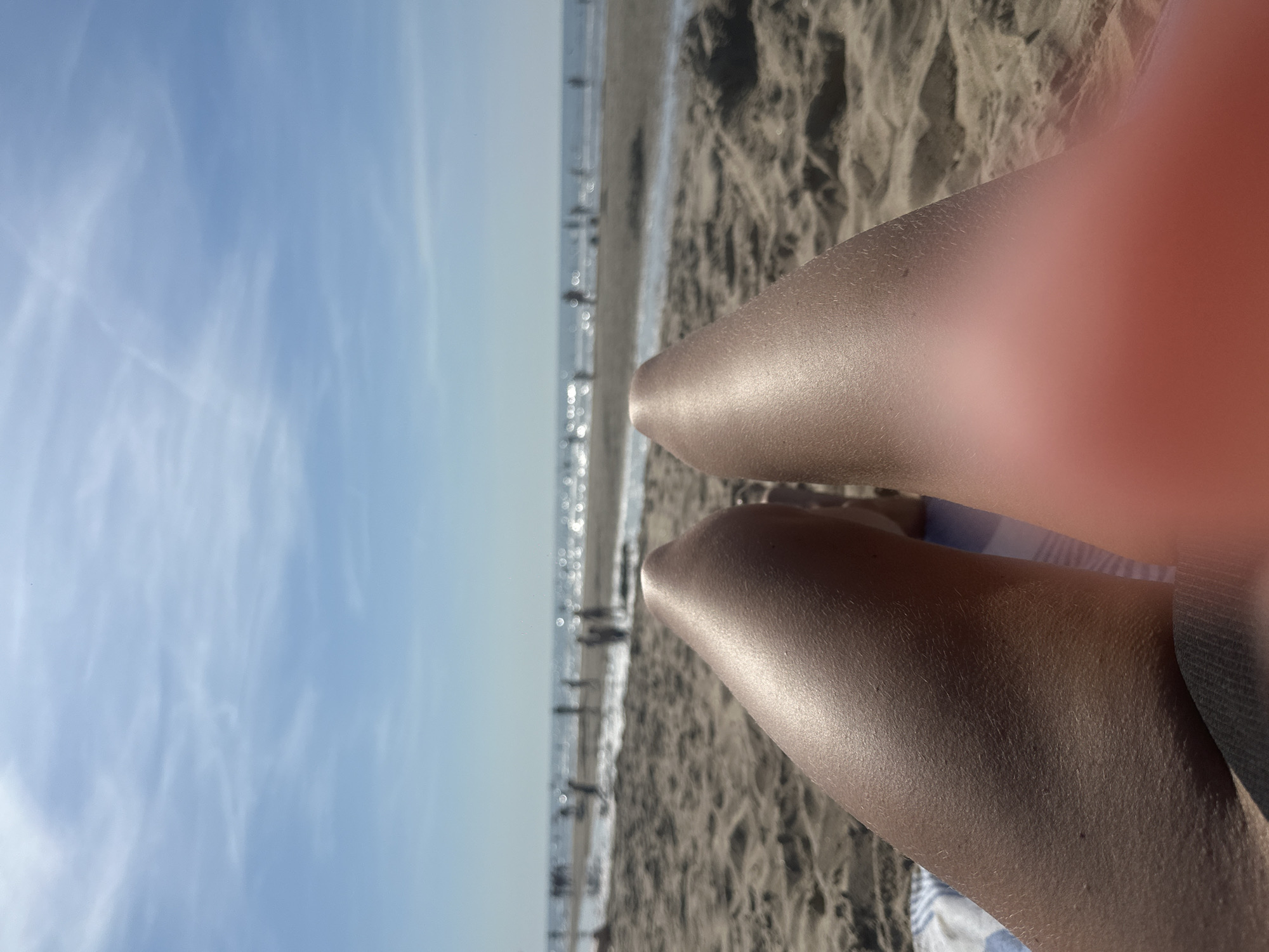 The image shows a first-person perspective of legs on a beach, with the ocean and sky in the background. The legs are tanned and appear to be resting on a beach towel. The beach itself is sandy, with some footprints and ripples in the sand. In the distance, there are several people scattered along the shoreline and in the water. The ocean is calm, with gentle waves breaking on the shore. The sky is blue with some scattered white clouds. The overall impression is one of a sunny day at the beach. This scene is consistent with a location in Zandvoort, Netherlands, a popular seaside resort town.