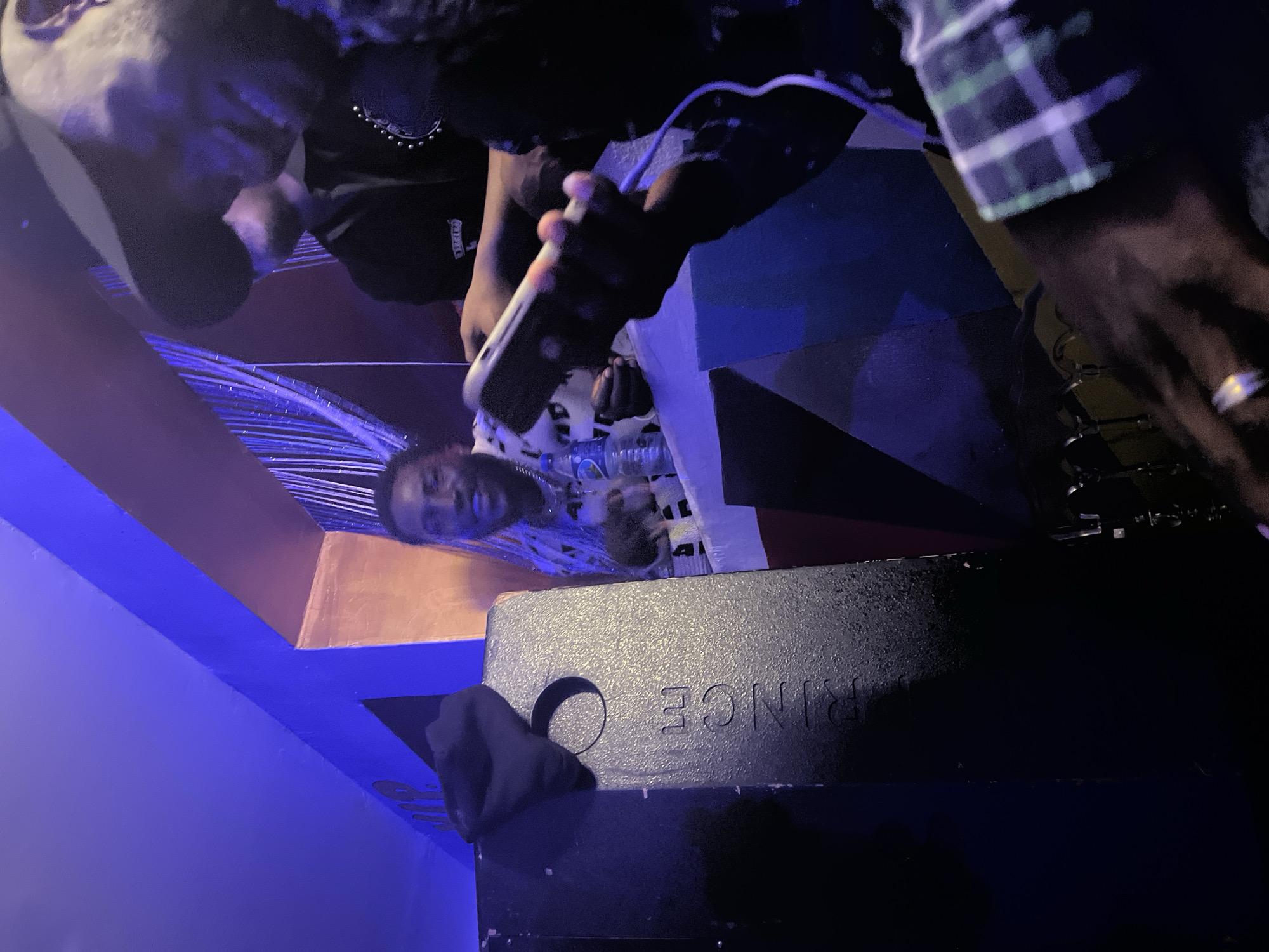 In Durumi, Nigeria, a dimly lit interior space, possibly a lounge or club, is captured with vibrant blue and orange lighting. Three individuals are present. On the left, a black speaker with the word "PRINCE" embossed on it is partially visible, with a black cloth draped over its top. In the middle background, a man with short, dark hair and a beard smiles, wearing a patterned shirt and a necklace. To his right, another person, mostly obscured, wears a dark cap and a dark jacket adorned with studs. In the foreground, a person's hands are visible, one holding a smartphone connected to a charger, and the other wearing a ring. The overall atmosphere appears social and relaxed.