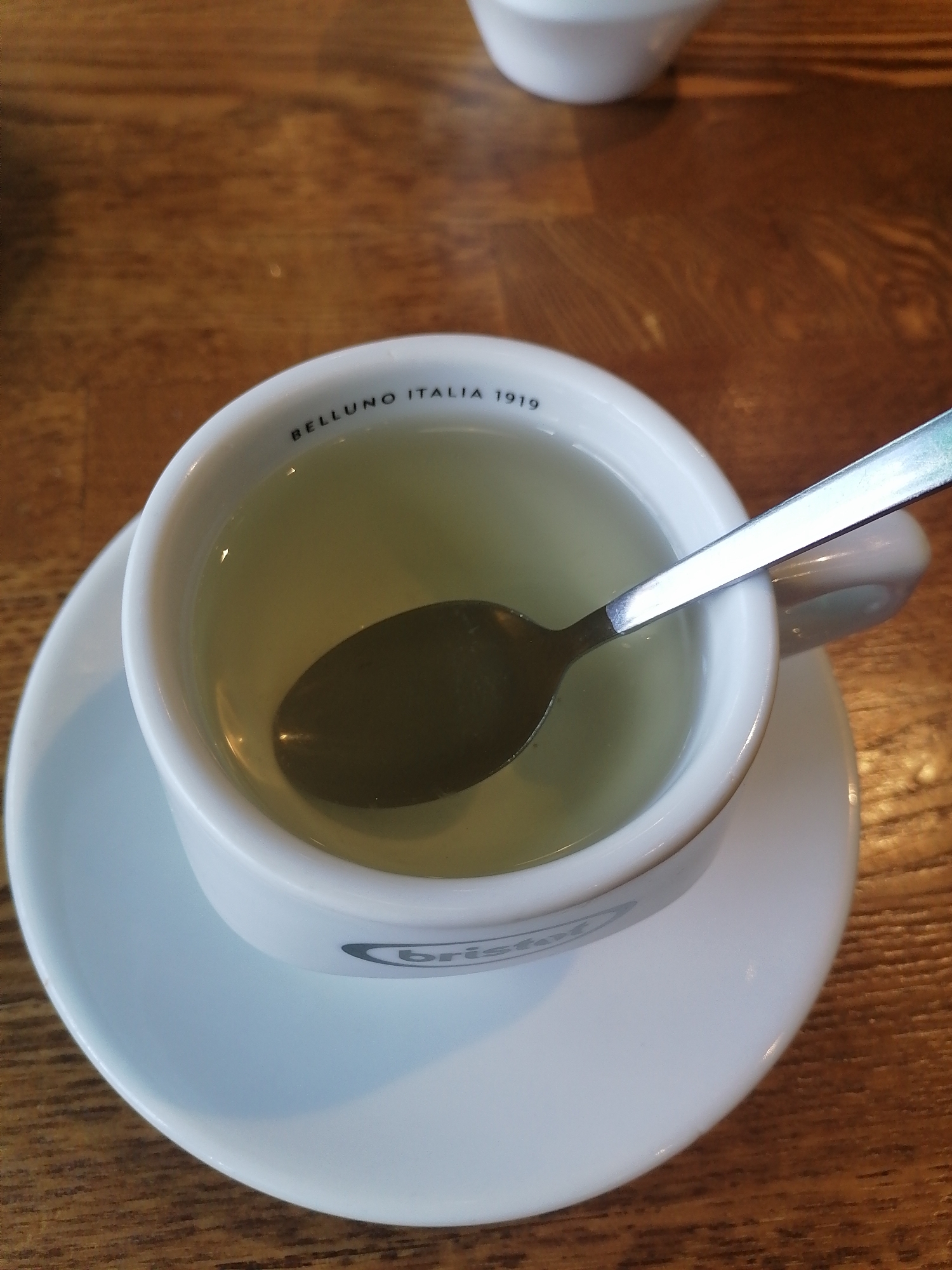 A close-up shot captures a white ceramic cup filled with a pale yellow liquid, likely tea, on a matching saucer. A silver spoon rests inside the cup, partially submerged in the liquid. The cup is adorned with black text that reads "BELLUNO ITALIA 1919" on the rim and a logo that appears to read "bristol" on the side. The objects are placed on a warm-toned wooden table. The lighting suggests an indoor setting, possibly a cafe or restaurant. There is no indication of the location being Moscow, Russia.