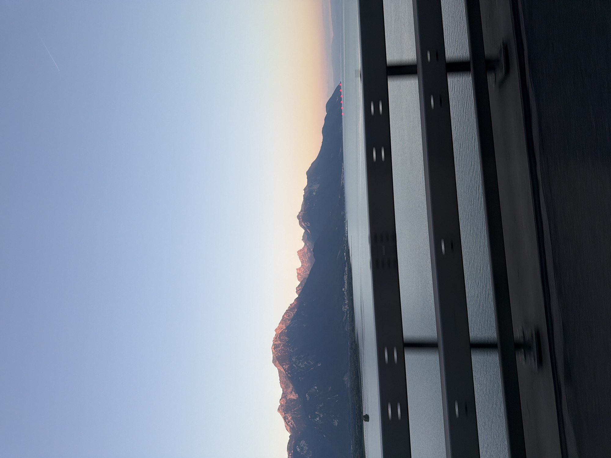 The image captures a panoramic view of the Alps and Lake Geneva, as seen from a bridge. The sky is a soft gradient of blue, transitioning to a pale orange at the horizon. A faint airplane contrail is visible in the upper right quadrant.
Dominating the middle ground are the majestic, snow-capped peaks of the Alps, bathed in the warm, rosy light of either sunrise or sunset. The lower slopes of the mountains are dark and shadowed, contrasting with the illuminated summits. A body of water, likely Lake Geneva, stretches across the lower half of the image, its surface calm and reflecting the light. Along the shoreline, a small town is nestled at the foot of the mountains. In the far right of the lake, a string of red lights can be seen, possibly marking buoys or a distant structure.
In the foreground, a dark metal guardrail runs horizontally across the frame, its bars and supports slightly blurred, suggesting movement as if captured from a moving vehicle. The road surface beneath the guardrail is also dark and shows motion blur.
The scene appears to be a scenic overlook, possibly part of a road or pathway constructed along the edge of Lake Geneva, offering a breathtaking vista of the natural landscape. The specific location within Villeneuve (VD), Switzerland, is suggested by the presence of the lake and the proximity to the Alps. The lighting indicates it is either early morning or late evening. There are no people or animals visible in the frame.