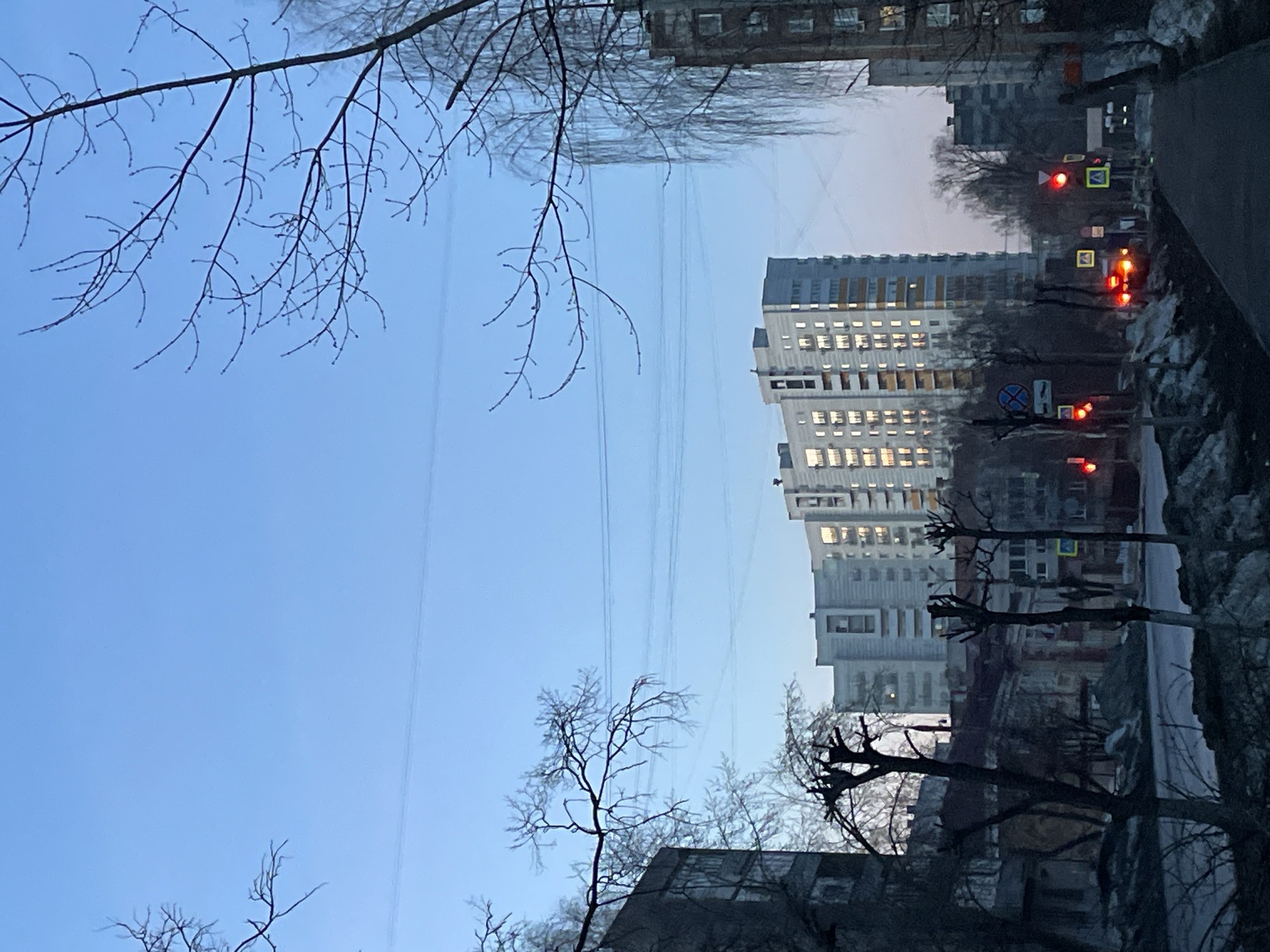 The image captures a street in what appears to be a residential district. A tall, multi-story building with numerous lit windows dominates the center of the frame, suggesting the presence of residents. Power lines stretch across the sky, adding to the urban environment. Along the street, there are barren trees, indicating a colder season, possibly late fall or winter. The presence of traffic lights, some showing red, suggests vehicular movement and urban traffic control. Street signs are also visible, further indicating a structured urban landscape. The overall setting suggests a typical urban scene, potentially located in a Russian city, considering the architectural style and environmental conditions. Based on the context provided, it is presumed this image is taken in Izhevsk, Russia.