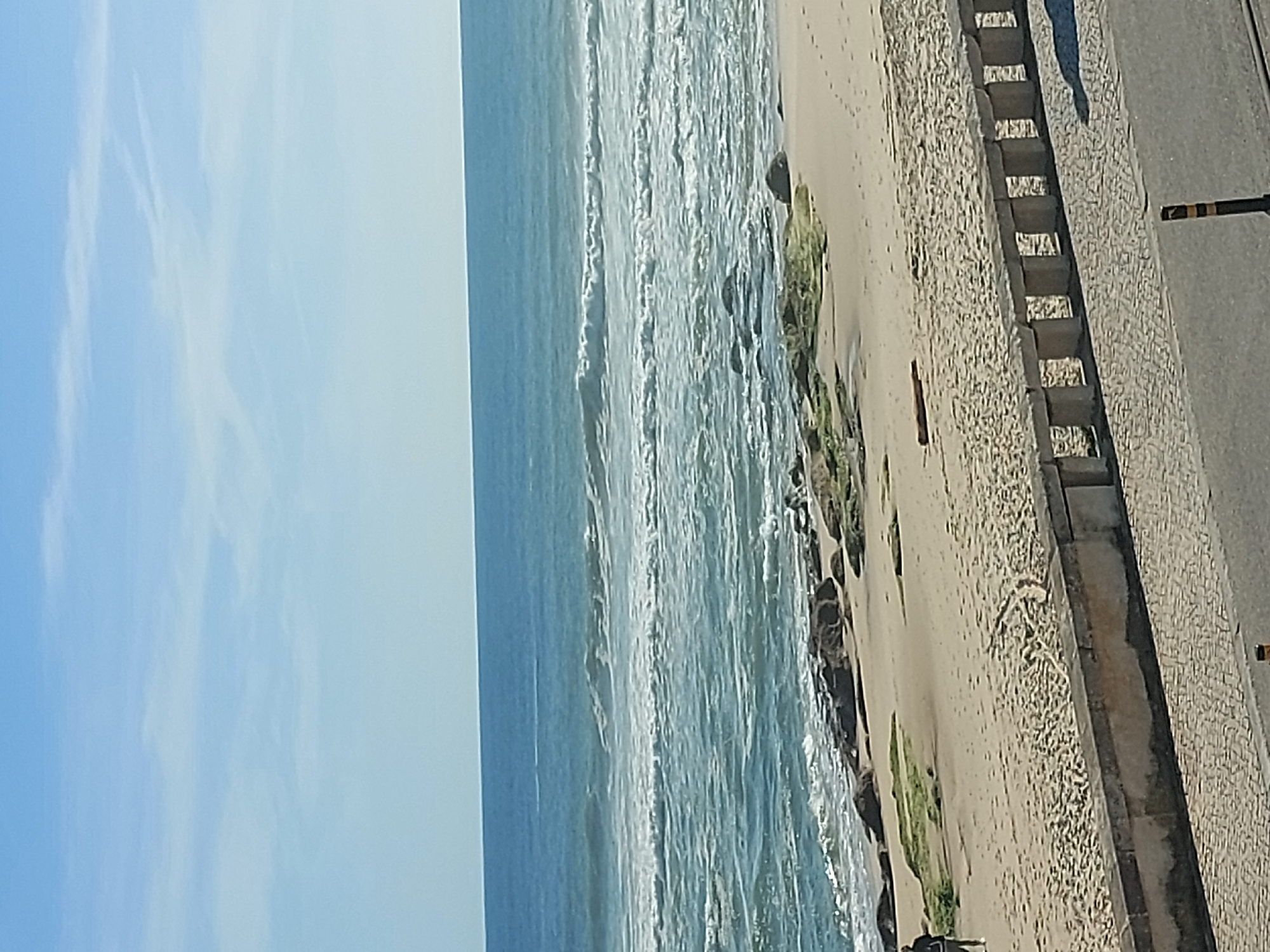 The image depicts a beach scene in Figueira da Foz, Portugal. The horizon is horizontal, with the ocean to the left. There are waves breaking on the beach, which is composed of sand. To the right of the beach is a stone seawall, separating the beach from an asphalt road. There are some steps built into the seawall. The sky is blue and clear. Some sparse vegetation is visible on the beach. A person is seen standing by the edge of the beach near the seawall.