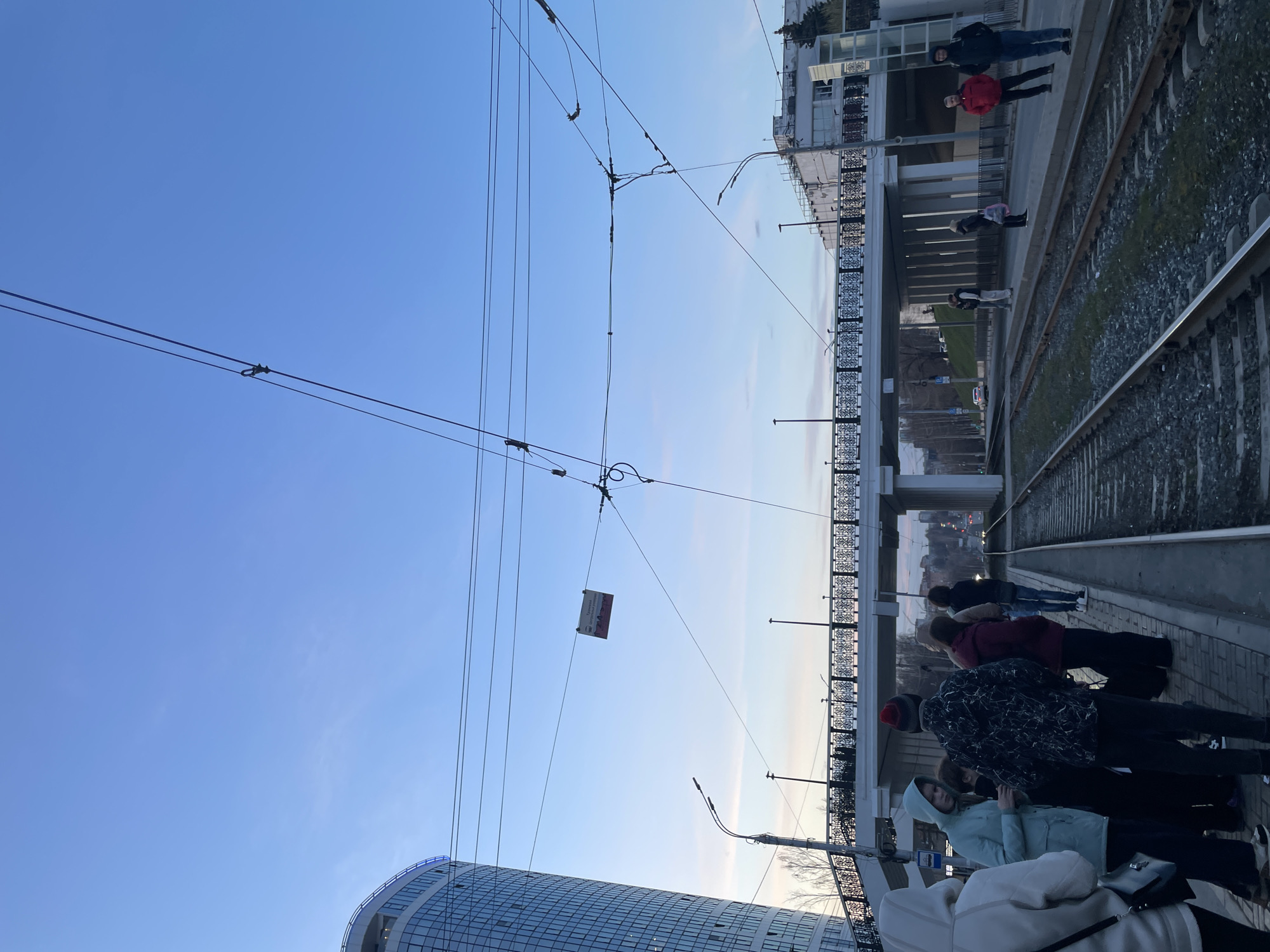 The image depicts an urban landscape in Izhevsk, Russia, likely at a tram stop or platform alongside railway tracks. The scene is illuminated by either early morning or late afternoon light, with a clear blue sky overhead that transitions to a lighter, hazy pale blue near the horizon.
In the foreground and midground, a group of approximately eight to ten people are gathered on a paved platform next to two sets of railway tracks. They appear to be waiting, possibly for a tram, and are dressed in casual attire, including jackets and hats, suggesting cool weather.
Above the platform and tracks, a concrete and metal bridge or overpass with ornate railings spans across the frame. A few additional people are visible on this bridge. Overhead, a dense network of tram or trolleybus wires crisscrosses the sky. Suspended from these wires is a rectangular white sign with red and black text. Partially legible words include "Изготовление" (Manufacturing/Production) and "Грузовая техника" (Cargo equipment), with some red graphical elements at the bottom.
To the far left, a modern, distinctive building with a curved glass facade and numerous windows occupies the lower portion of the image. Another building is visible beyond the bridge on the right. Several street lamps are also present throughout the scene, contributing to the urban setting.