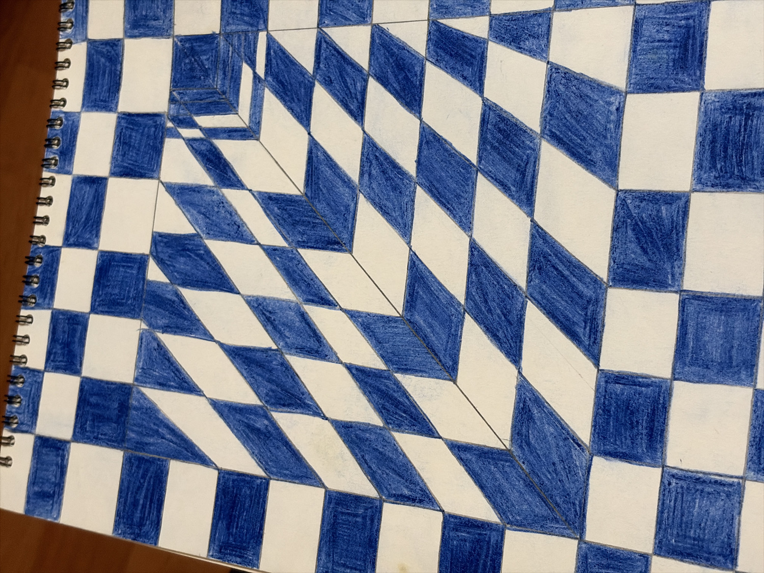 The image displays an open spiral-bound sketchbook, largely filled with an abstract drawing. The drawing features an optical illusion composed of a checkerboard pattern of blue and white squares and rhombuses. This geometric pattern creates a strong sense of three-dimensionality, appearing to recede into a central, cubic void or "hole" primarily located in the upper-left half of the drawing.
The blue sections are hand-colored with what appears to be crayon or colored pencil, showing visible strokes and texture. The white sections are the untouched paper. Faint pencil lines are discernible, indicating the underlying grid used to construct the illusion. The pattern transitions from a more conventional, flatter checkerboard along the outer edges of the drawing to a highly angled, perspective-driven grid in the central area, enhancing the illusion of depth.
The sketchbook has a black spiral binding visible along its left edge. A warm, brown surface, possibly a wooden table, is partially visible beneath the sketchbook at the bottom and left. The lighting is even, illuminating the artwork without casting harsh shadows. There are no people or discernible text in the image. The artwork evokes the style of Op Art, playing with visual perception and geometric abstraction.