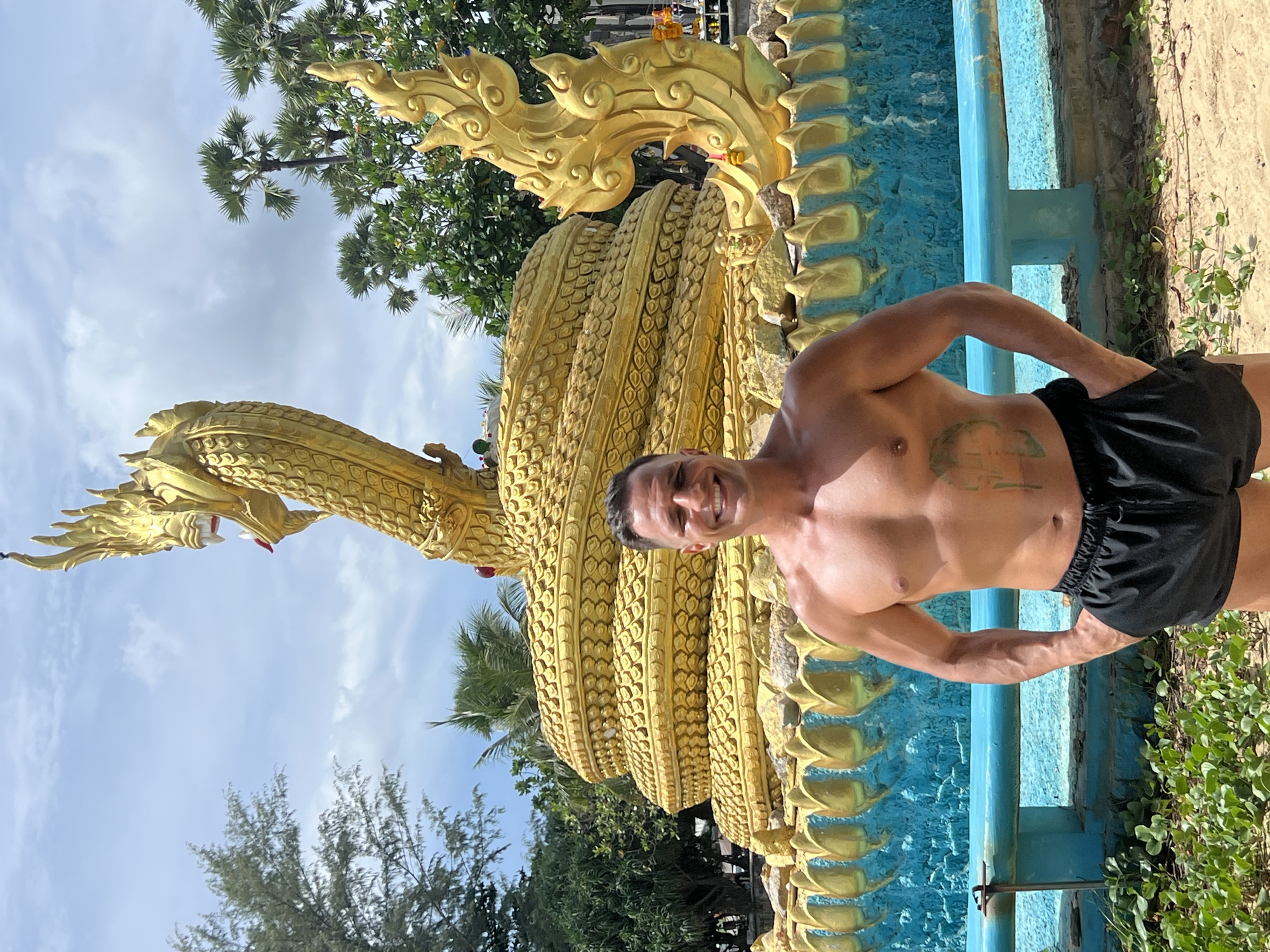 A shirtless man with black shorts stands on a sandy beach in front of a large golden serpent statue, a landmark in Karon, Thailand. The statue, with its coiled body and intricate scales, is partially surrounded by a turquoise blue base adorned with golden decorative elements. The man is smiling directly at the camera, with his hands in his pockets. Lush green foliage and palm trees are visible in the background, under a bright, partly cloudy sky.
