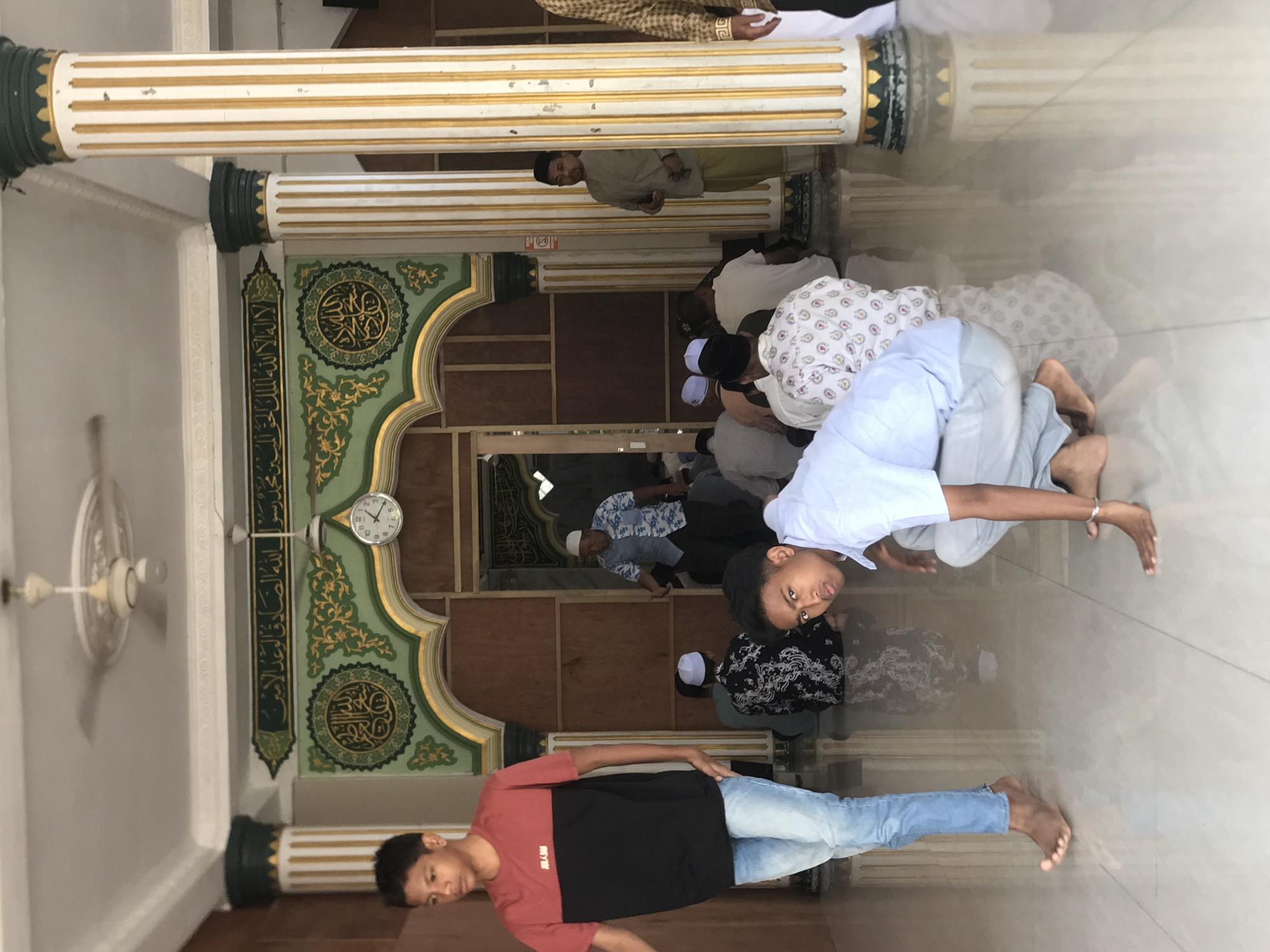 The photo shows the interior of the Islamic Center Lhokseumawe mosque in Lhokseumawe, Indonesia. Several people are praying. One person is standing in the foreground, and another is kneeling in prayer in the mid-ground. Architectural details include pillars, ornate green and gold wall decorations with Islamic calligraphy, a clock on the wall, and a ceiling fan. The floor appears to be tiled.