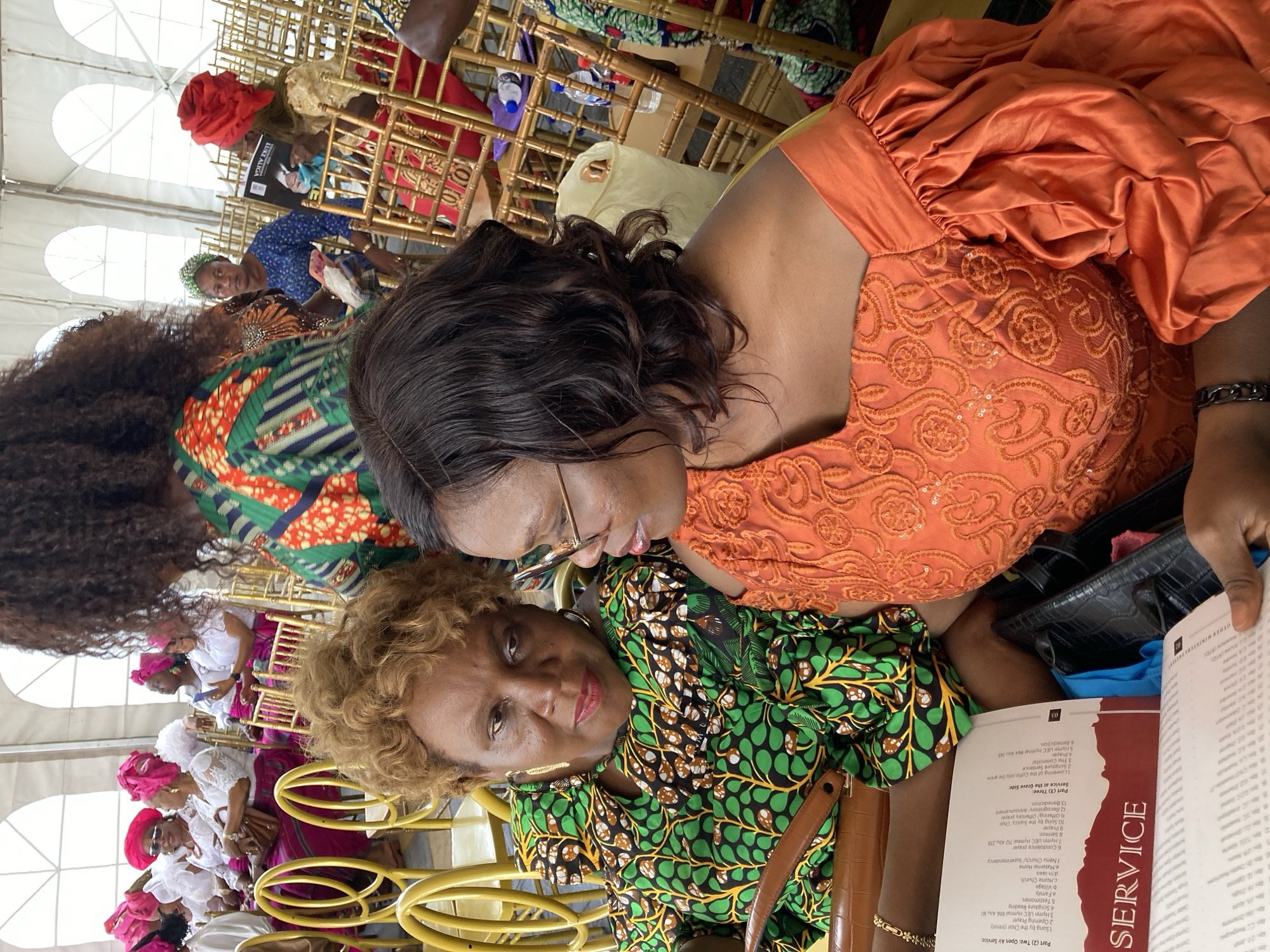 Two middle-aged women are seated in a chair and looking at a service booklet. One woman is wearing an orange dress with puffed sleeves, and the other is wearing a green patterned dress. They are attending a service at "Nnenu Church/Superintendency" as it is stated in the booklet and in the city of Nnenu, Nigeria. The booklet is open to a page titled "SERVICE". The booklet includes the hymns and prayers for the service, one of which is "Hymn UEC Hymnal 712 Abu 239". Other people are seated behind them in gold-colored chairs, and a large white tent is overhead. One of the other people in the background wears a red head covering.
