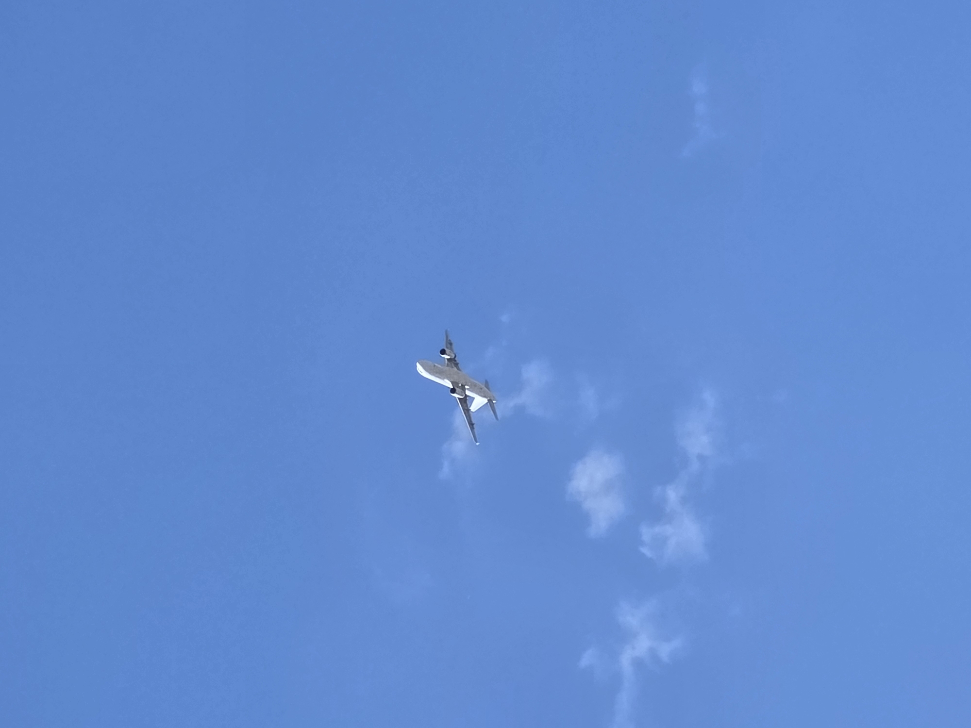 A silver jet aircraft is flying in a clear blue sky. Several scattered clouds are visible. The location cannot be determined as Varna, Bulgaria with the information present in the image.