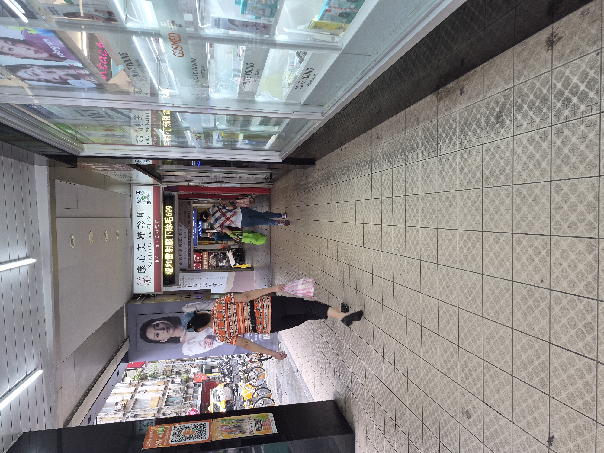 The image depicts a street scene in Taipei, Taiwan, specifically in an area with covered walkways adjacent to shops. The perspective is from ground level, looking down a tiled walkway. Two women are walking away from the camera, one in the foreground and another further down the walkway. The woman in the foreground is wearing an orange and white striped shirt and black pants, carrying a pink plastic bag. The woman behind her is wearing a checkered shirt and blue jeans, carrying a green reusable shopping bag.
On the left side of the walkway, there are posters and advertisements. One prominent poster features a portrait of a woman. Further down, there are bicycles parked, and a yellow taxi is visible. On the right side of the walkway, there is a storefront with large glass windows displaying various cosmetic products. The storefront appears to be a beauty clinic or a retail store selling beauty products, with prominent signage like "OLIVE YOUNG" and "COSMED" visible. The environment suggests a commercial district with pedestrian traffic. The tiled ground has a repeating pattern, and some areas show signs of wear or dirt. The overall lighting suggests it is daytime.