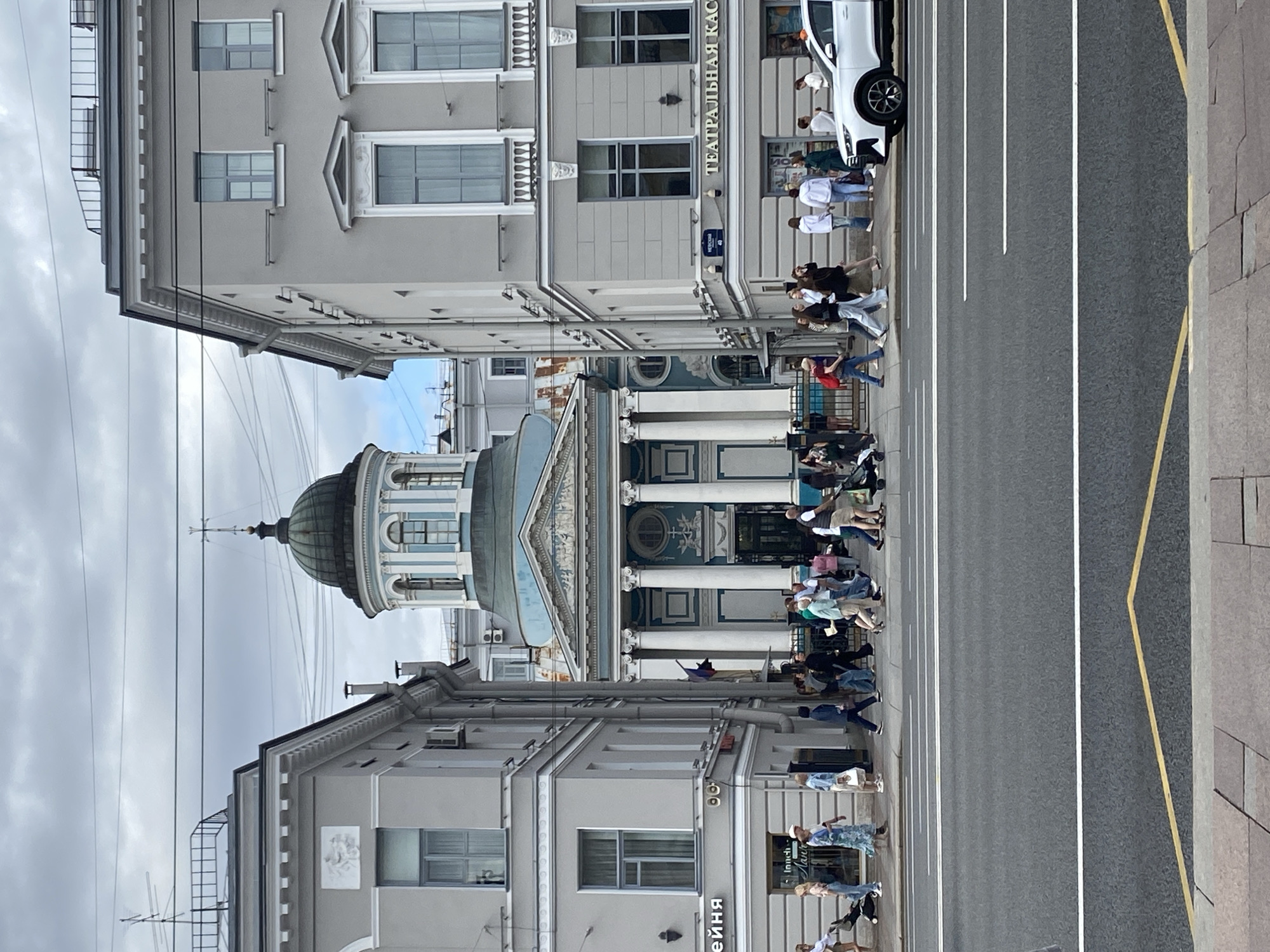 The image captures a street scene in Saint Petersburg, Russia, with a prominent domed building in the background that appears to be a church or religious institution. The architecture suggests a classical or neoclassical style, with columns and ornate detailing. In the foreground, a wide asphalt road is visible with white lane markings. Several people are walking on the sidewalk to the right of the road, some pushing strollers. A white car is parked on the right side of the street. The buildings flanking the street are multi-story, gray structures with large windows. The sky is overcast, indicating a cloudy day. The overall impression is of a busy urban environment.