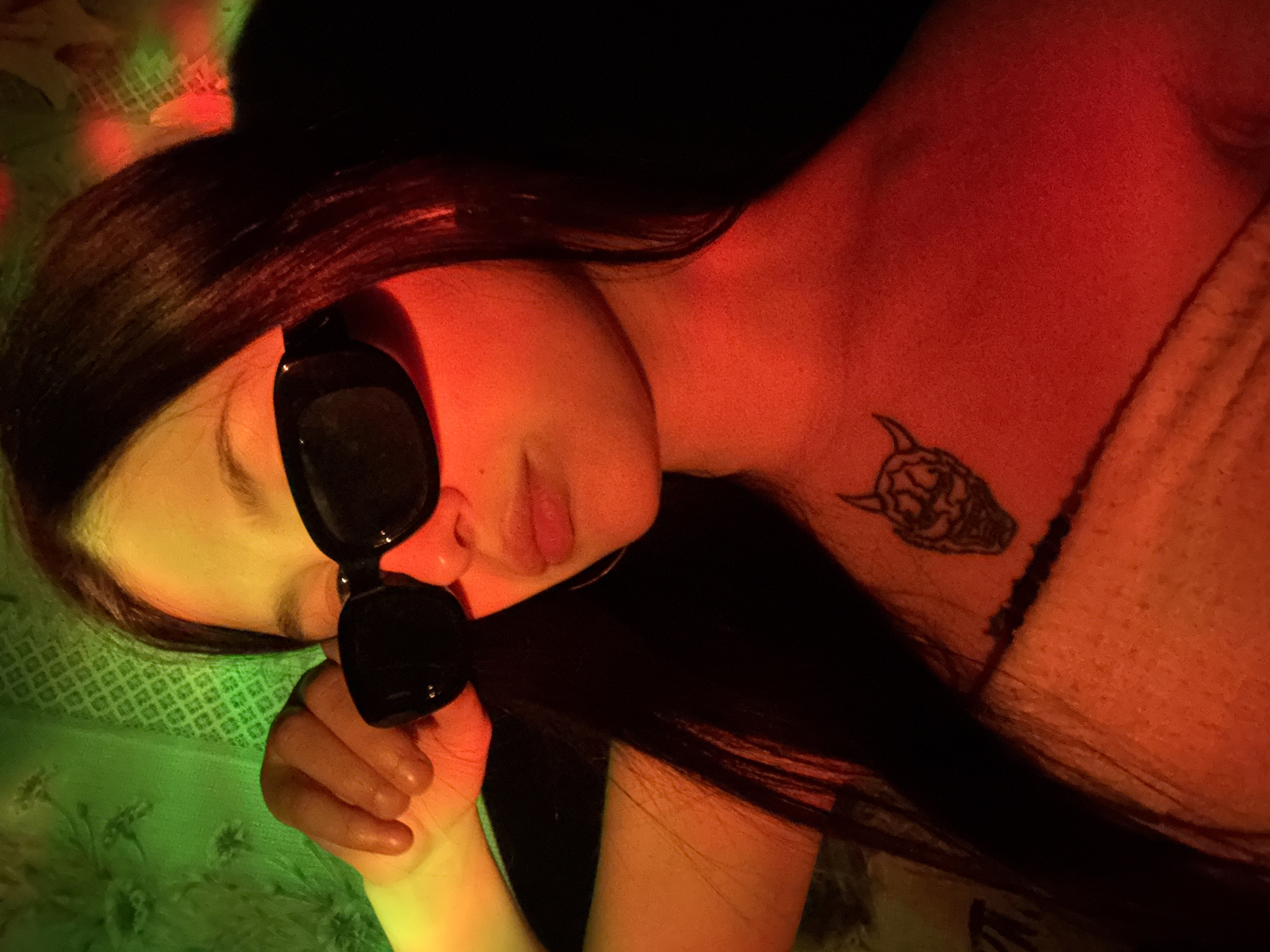 A person is lying down. The person has long dark hair and is wearing sunglasses. There is a tattoo on the person's neck. The background has green and red lighting. The city is not mentioned nor can it be confirmed by the image.