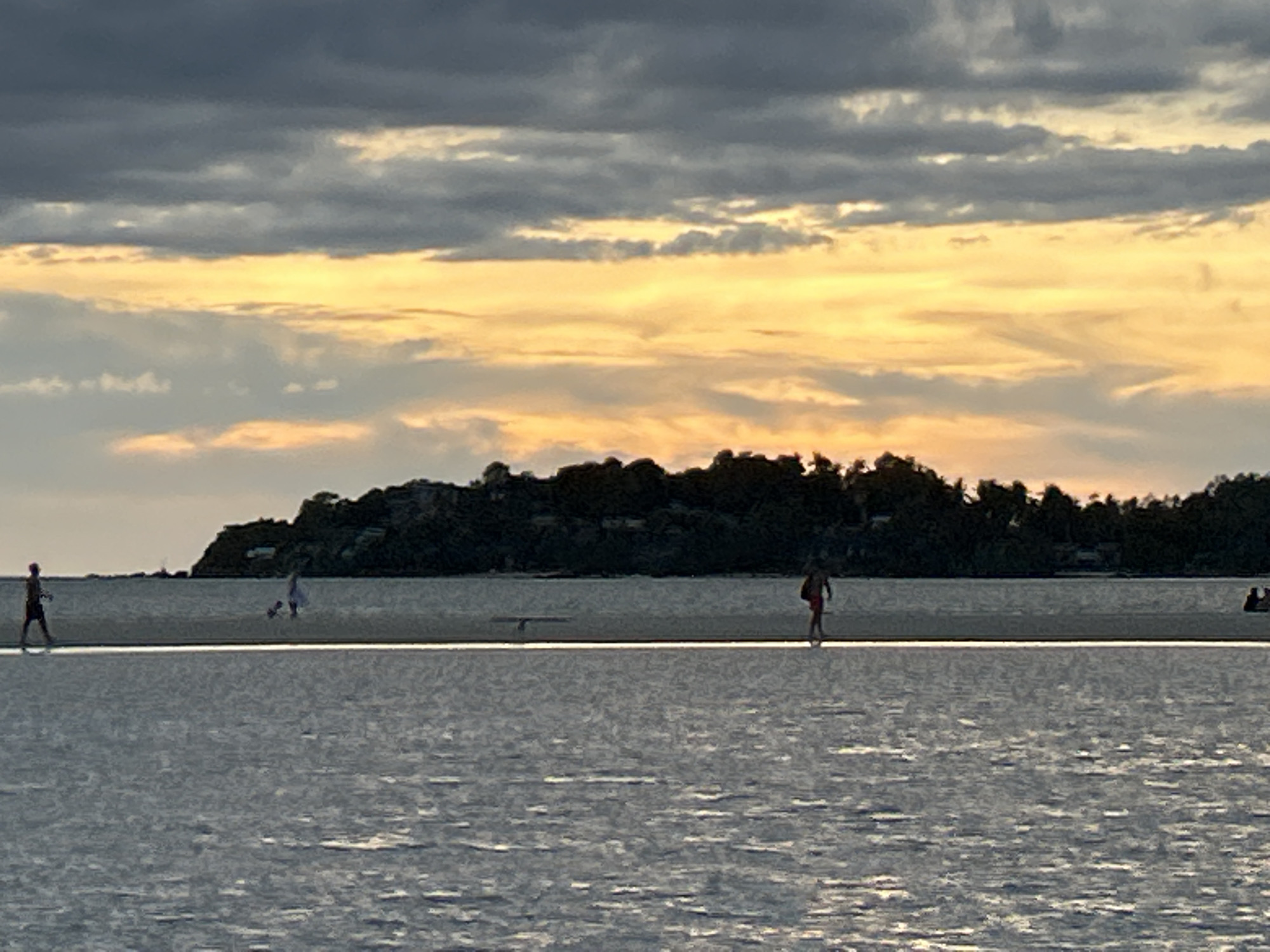 The image depicts a coastal scene at sunset in ตำบลเกาะพะงัน, Thailand. The foreground is dominated by water with subtle wave patterns. A narrow sandbar stretches across the middle ground, connecting to an island densely covered with trees. Several people are walking on the sandbar, appearing as silhouettes. In the background, the sky is a mix of gray clouds and orange hues from the setting sun. The island's silhouette is dark against the bright sky.