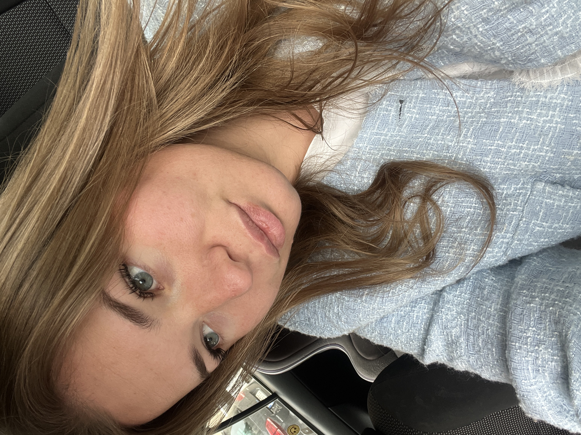 A young woman with light brown hair, blue eyes, and fair skin is in the passenger seat of a vehicle. She is wearing a light blue jacket with a textured fabric. The vehicle appears to be stationary, as evidenced by the visible surroundings outside the window. A reflection in the window of a building with signage suggests that the location is the city of Budva, Montenegro. The interior of the vehicle is visible, including the dashboard and part of the center console.