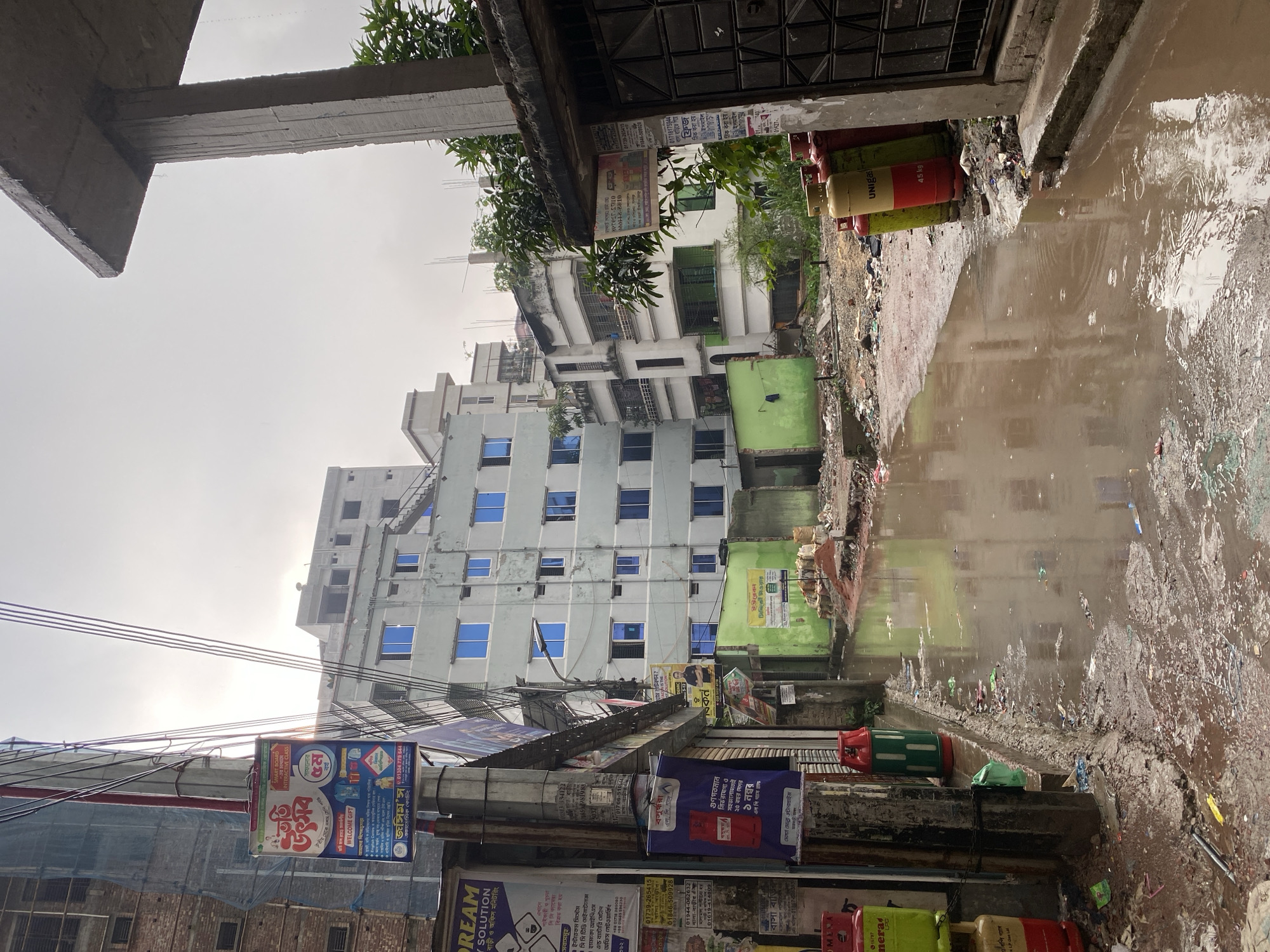 The scene depicts a flooded street in Dhaka, Bangladesh, reflected in a large puddle filled with debris. The street is lined with buildings, some tall and modern, others older and in disrepair, showing a mix of architectural styles. Utility poles are cluttered with various promotional banners and posters advertising services like coaching centers, security systems, and gas companies. Several gas cylinders are visible near the flooded area. The sky is overcast and the general atmosphere suggests recent or ongoing rainfall. The overall impression is a congested, urban environment struggling with drainage issues after a rain event.