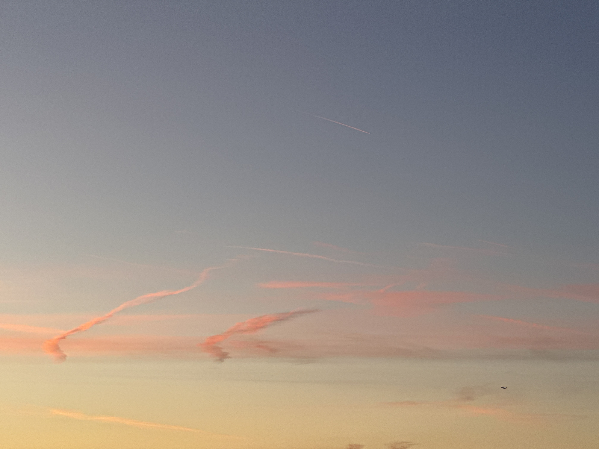 This is a vertical, medium-shot photograph of a sky at sunset, with airplane contrails and two birds visible. The sky transitions from a pale blue at the top to a soft yellow-orange at the bottom. There is a faint airplane contrail, tinged with pink, extending diagonally from the upper right to the center left of the frame. Below this, more prominent contrails, painted in shades of orange and pink by the setting sun, stretch across the middle of the image. In the lower right quadrant of the image, two small dark silhouettes of birds are in flight. The scene is devoid of any structures or human presence.