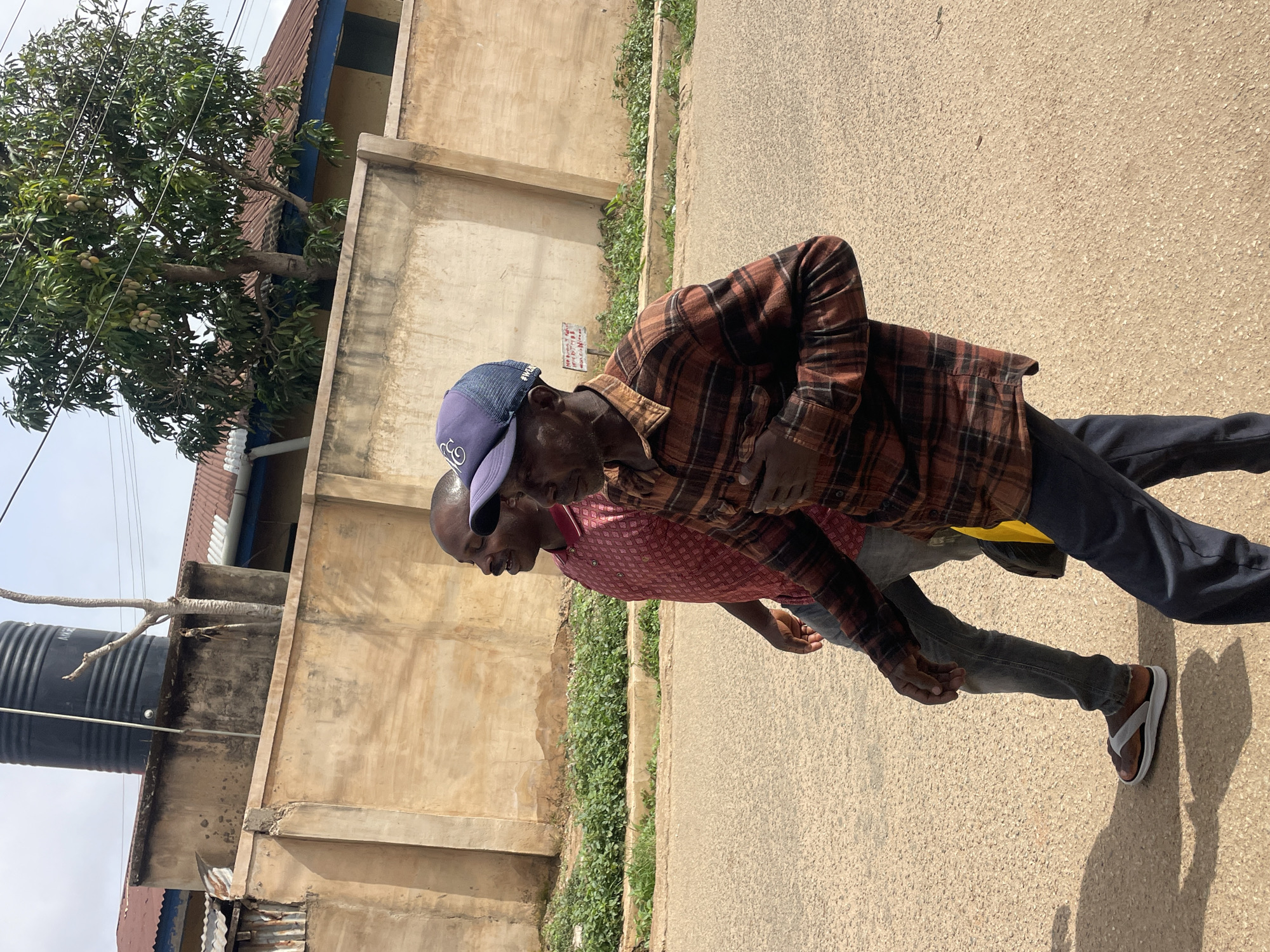 Two men walk along a paved road in Jos, Nigeria. The first man, closer to the camera, wears a brown and black plaid shirt, dark pants, and brown sandals. He has a blue baseball cap on. The second man wears a pink patterned shirt and jeans. A tall concrete wall runs alongside the road, with greenery growing at its base. A tree with green leaves is visible behind the wall. A black water tank and a partially visible building are in the background. The weather is sunny.