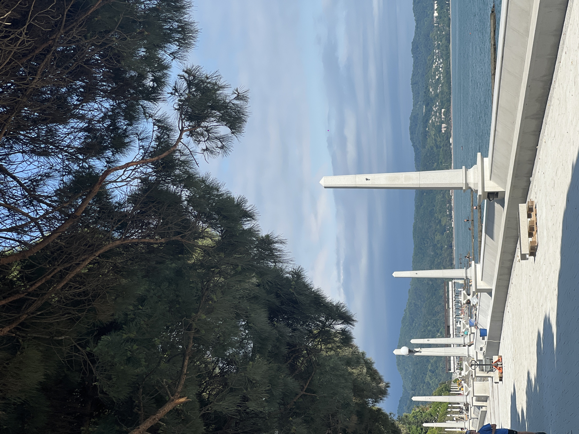 The image depicts a waterfront promenade with a paved walkway in the foreground, lined with decorative white obelisks and lampposts. A man in a blue shirt and dark shorts is jogging on the left side of the walkway. Beyond the promenade, the sea is visible, with a coastline featuring green hills dotted with buildings in the background. The sky is partly cloudy. The scene appears to be in a resort area, with some ongoing construction or maintenance work on the promenade, evidenced by pallets of materials. The overall atmosphere is bright and sunny, with a hint of the sea breeze.
